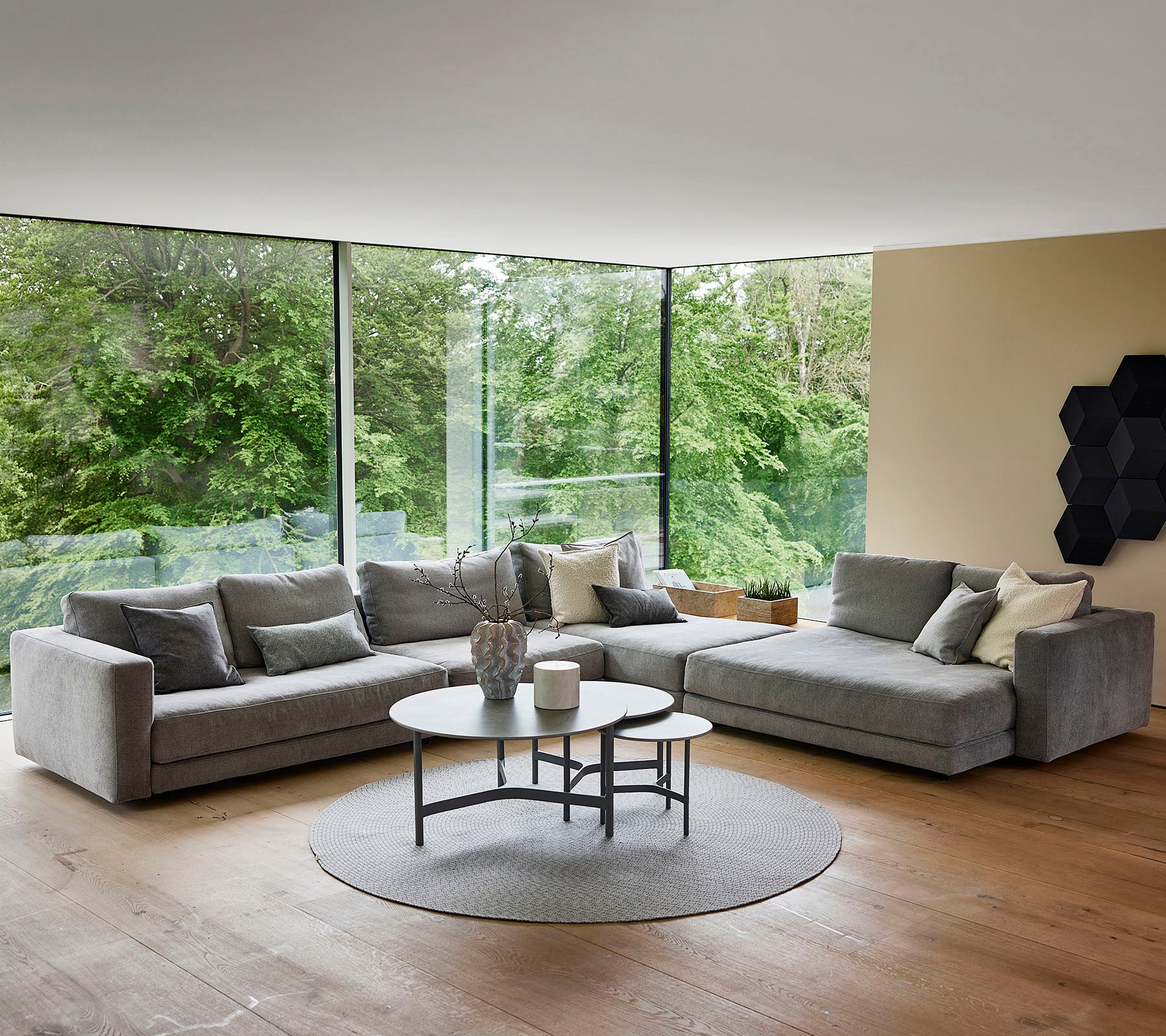 Un salon moderne et élégant avec un canapé d'angle en velours gris, une table basse ronde et de grandes fenêtres qui laissent voir la verdure à l'extérieur.