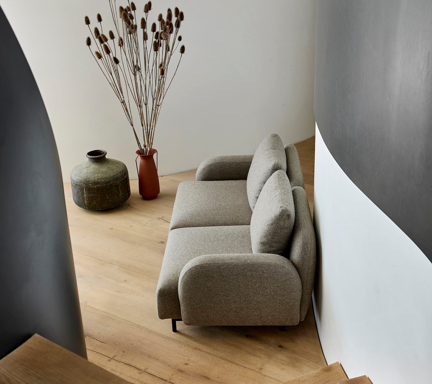 Un canapé beige confortable positionné à côté d'un vase décoratif et de plantes séchées, contre un mur courbé et un sol en bois.