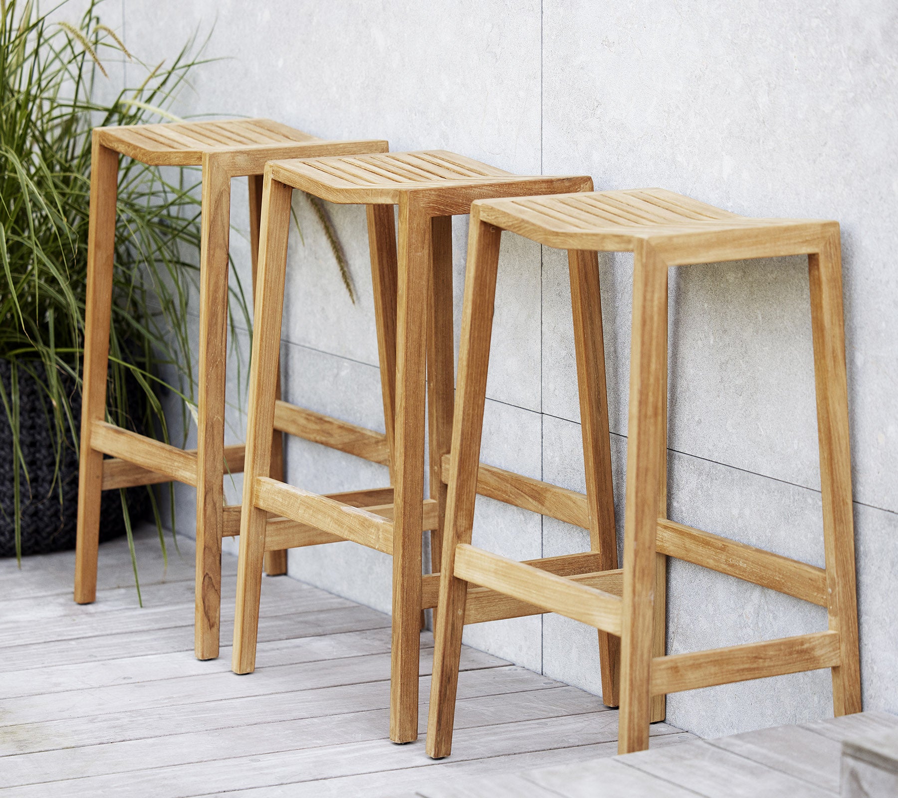 Trois tabourets en bois à bascule avec des sièges à lattes, présentant un design minimaliste et des lignes épurées, positionnés à côté d'une plante décorative.