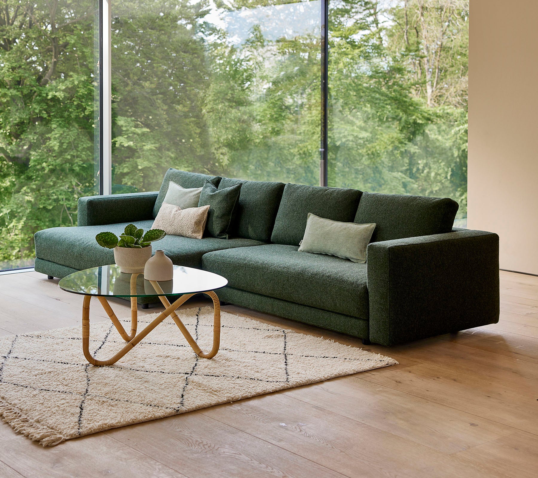 Un canapé vert moderne avec des coussins, accompagné d'une table basse ronde et d'un tapis décoratif, sur fond de verdure.