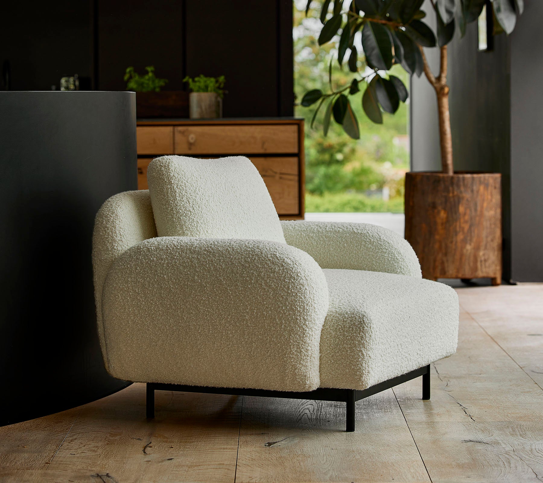 Fauteuil blanc cosy avec accoudoirs arrondis et une texture moelleuse, planté devant un arrière-plan de verdure et de mobilier moderne.