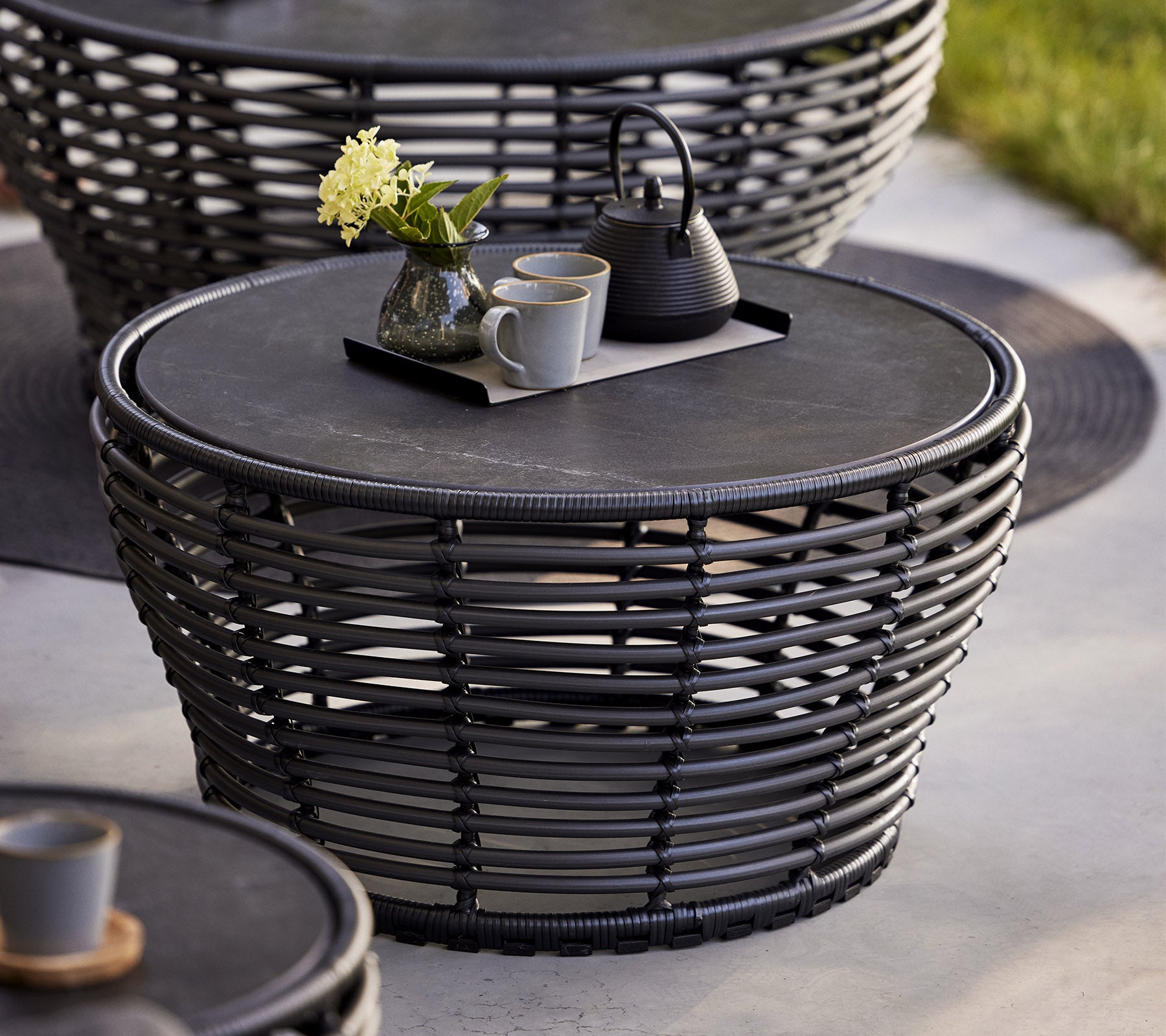 Table basse tissée élégante avec un dessus rond, comprenant un plateau décoratif avec une théière et des tasses, complété par un vase à fleurs.