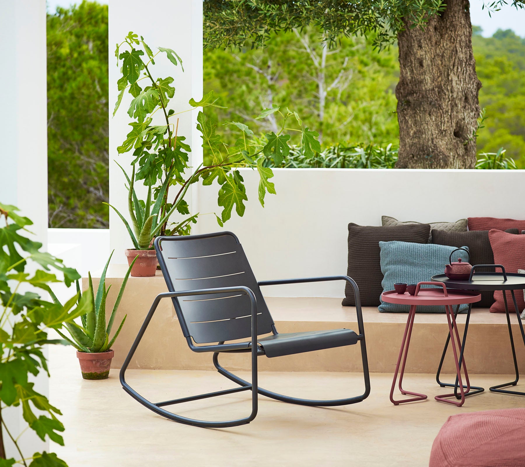 Chaise berçante élégante à côté de plantes en pot et de coussins, créant un coin salon cosy et accueillant avec une vue sereine.