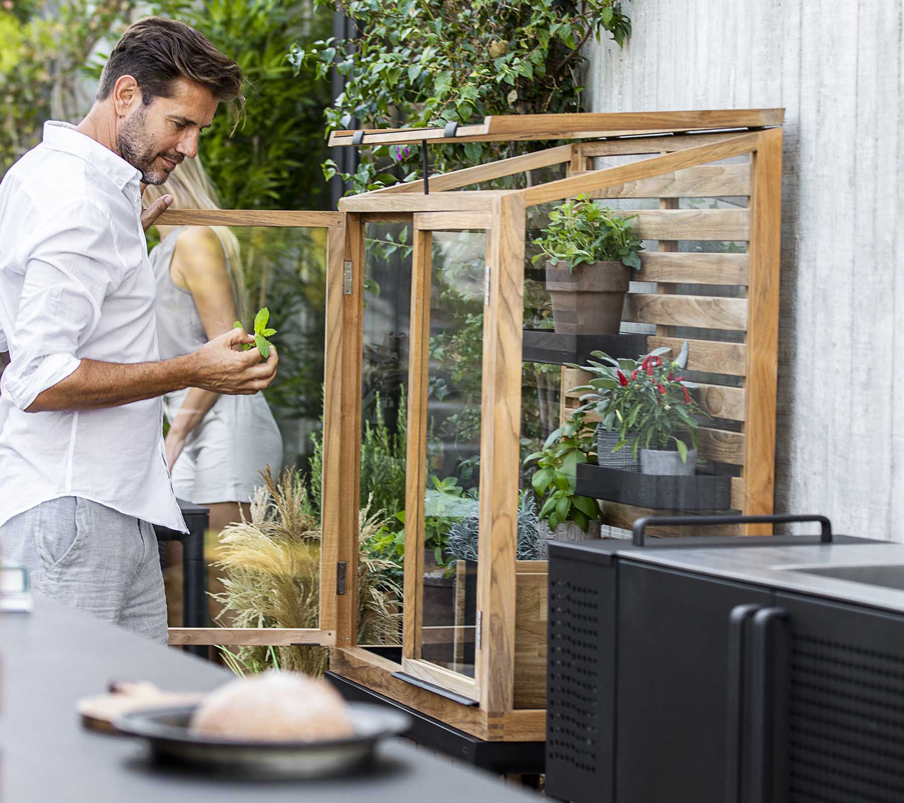 Homme s'occupant des plantes dans une unité d'exposition en verre, entouré de verdure, avec une cuisine moderne en premier plan.