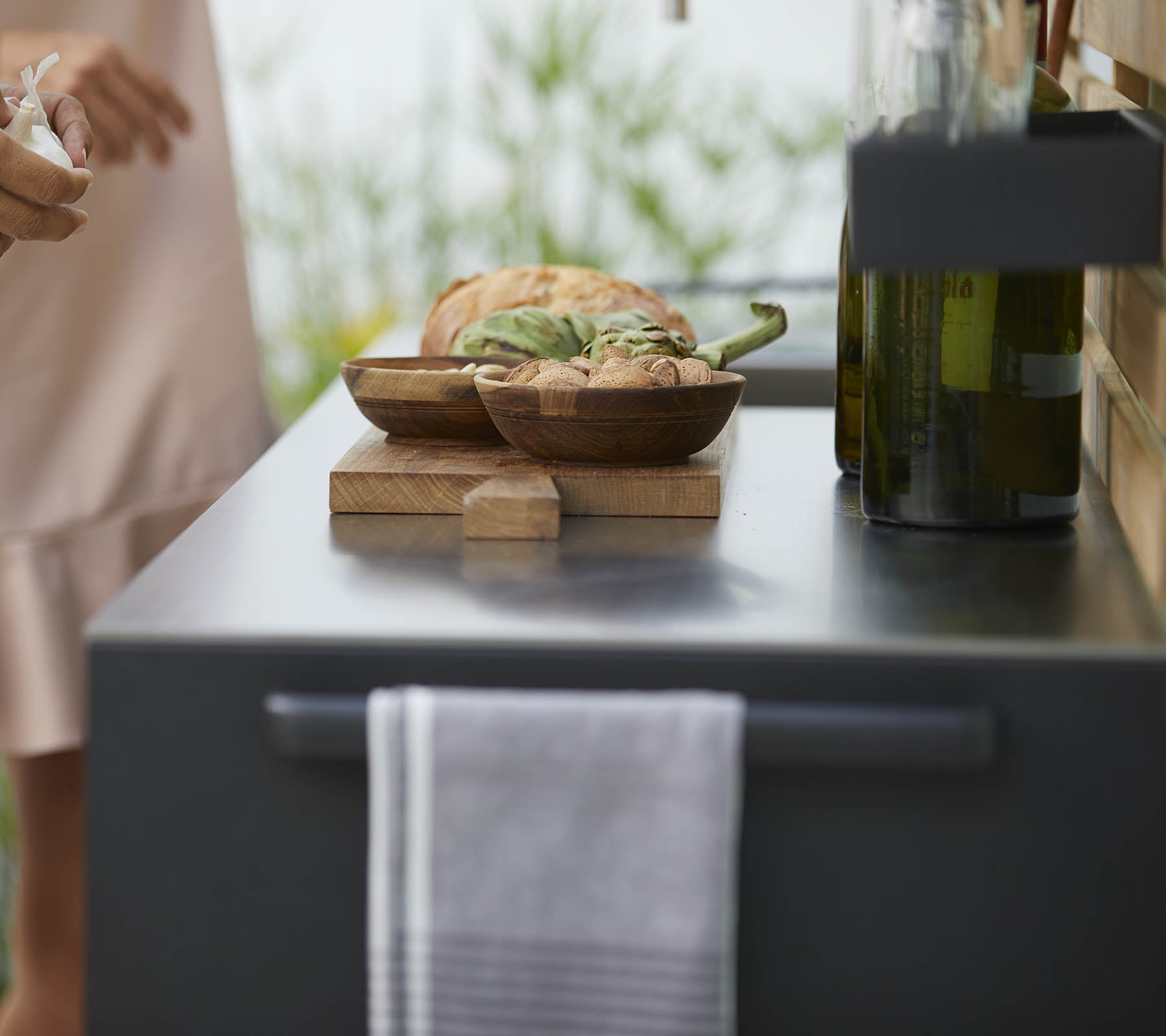 Un gros plan d'un plan de travail de cuisine avec des bols en bois de nourriture, une serviette et des bouteilles d'huile d'olive, avec des plantes douces en arrière-plan.