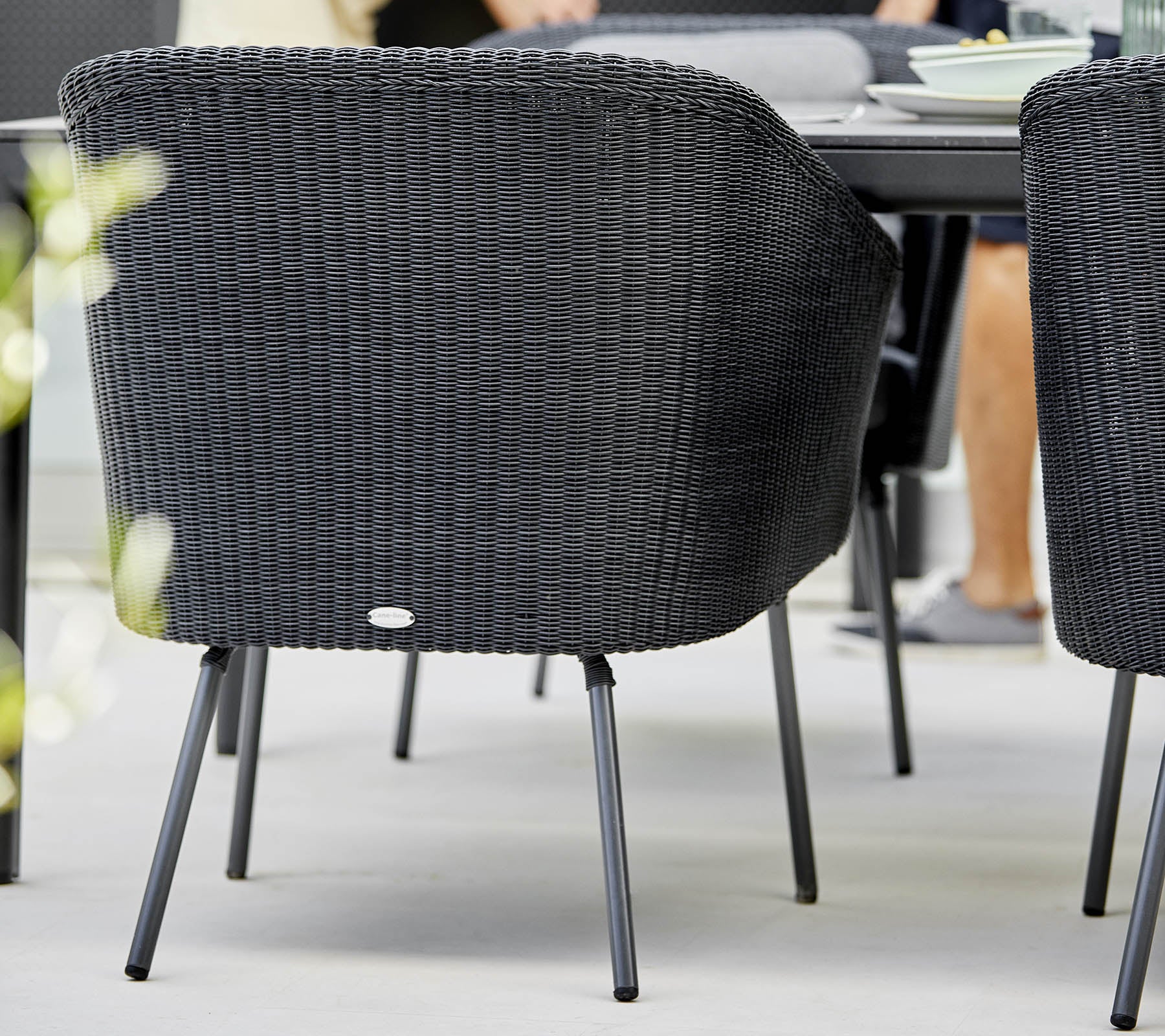 Chaise tissée noire avec un dossier incurvé et des pieds en métal, positionnée près d'une table à manger avec un flou doux sur les éléments environnants.