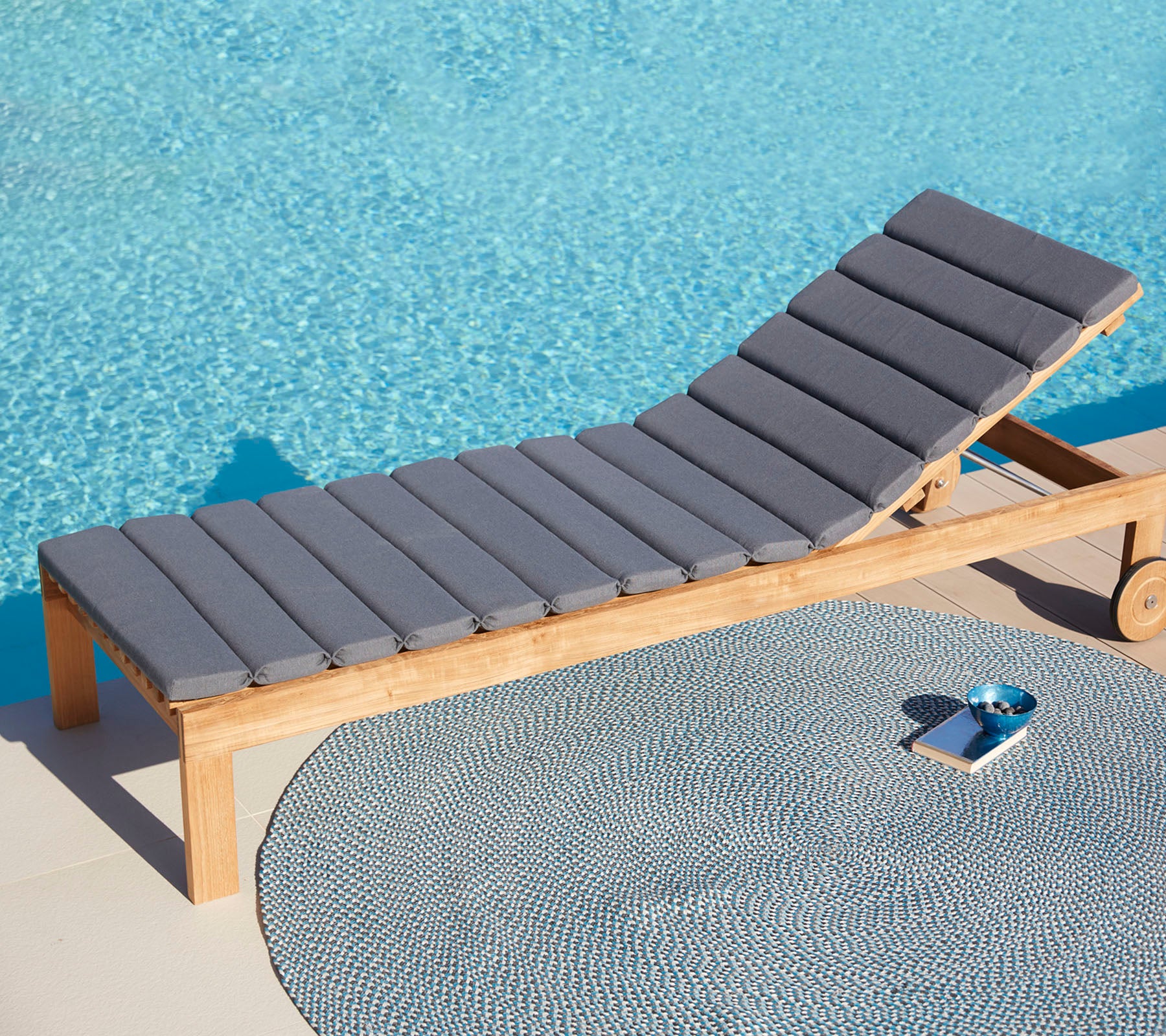 Chaise longue grise confortable à côté d'un tapis circulaire et d'un petit plateau, avec une surface d'eau bleue tranquille en arrière-plan.