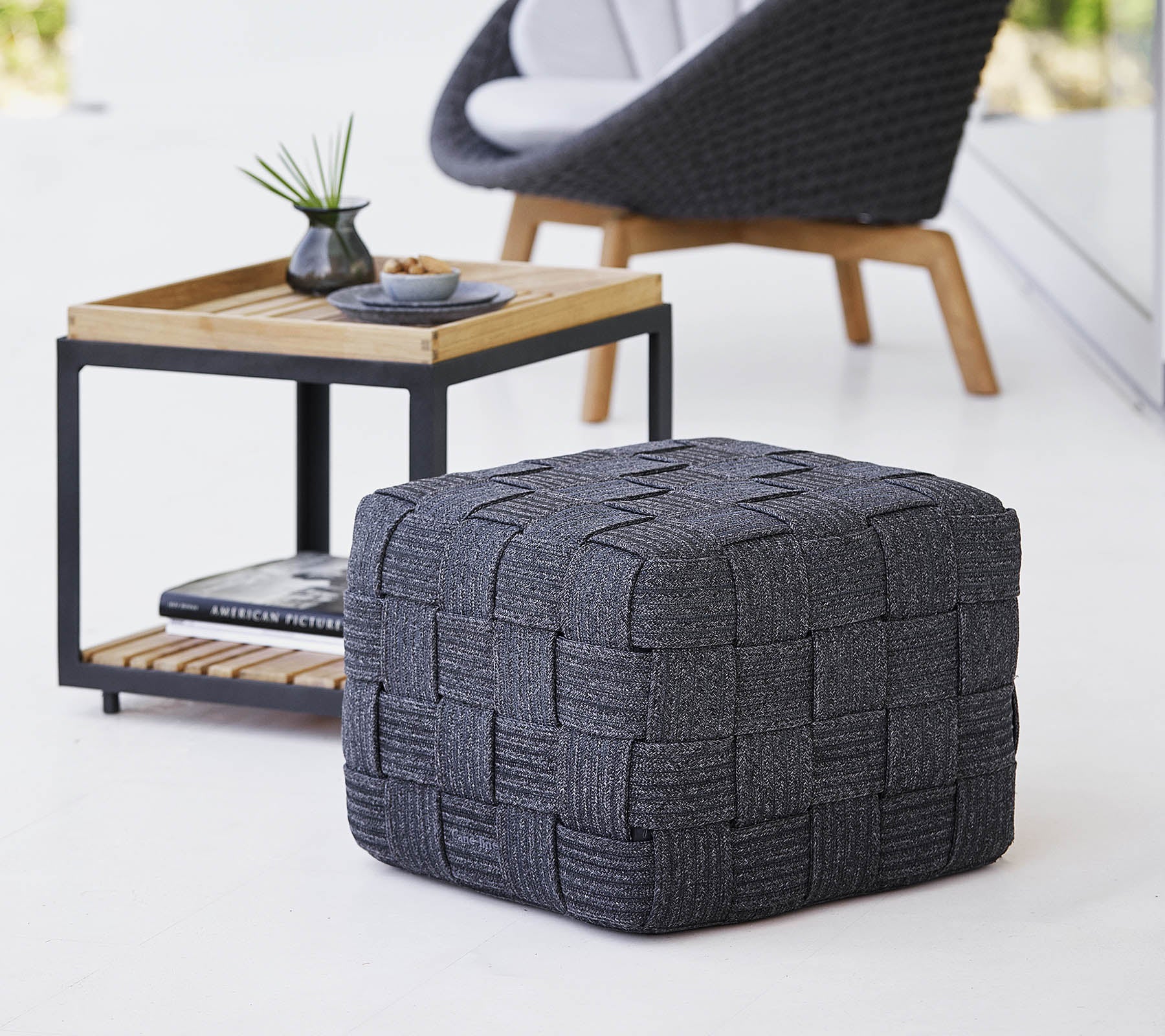 Un pouf tissé gris texturé situé près d'une table d'appoint en bois au design minimaliste, avec de petits objets décoratifs.