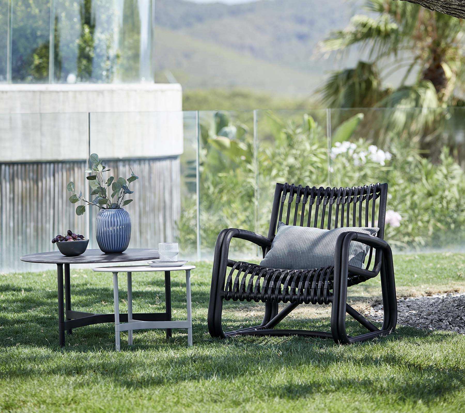 Chaise longue contemporaine avec un design à motifs, accompagnée d'une table d'appoint portant un vase et une plante en pot, placée dans un paysage naturel.