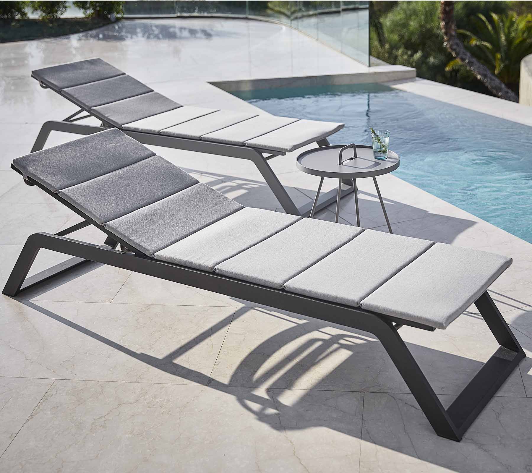 Deux chaises longues élégantes avec des coussins gris sont placées à côté d'une piscine, accompagnées d'une petite table d'appoint avec une boisson.