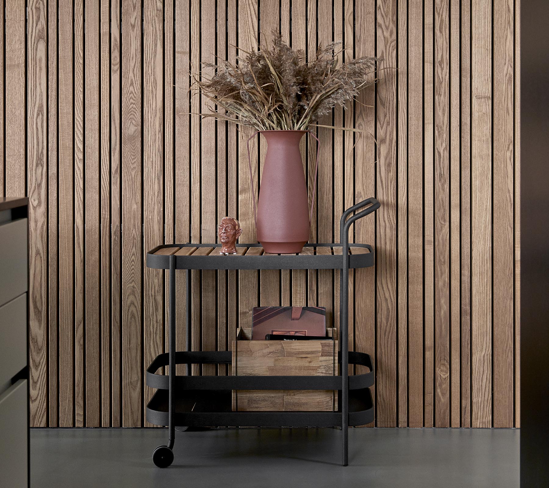 Un chariot élégant avec une finition sombre, doté de boîtes de rangement en bois et d'un vase rose avec des fleurs séchées.