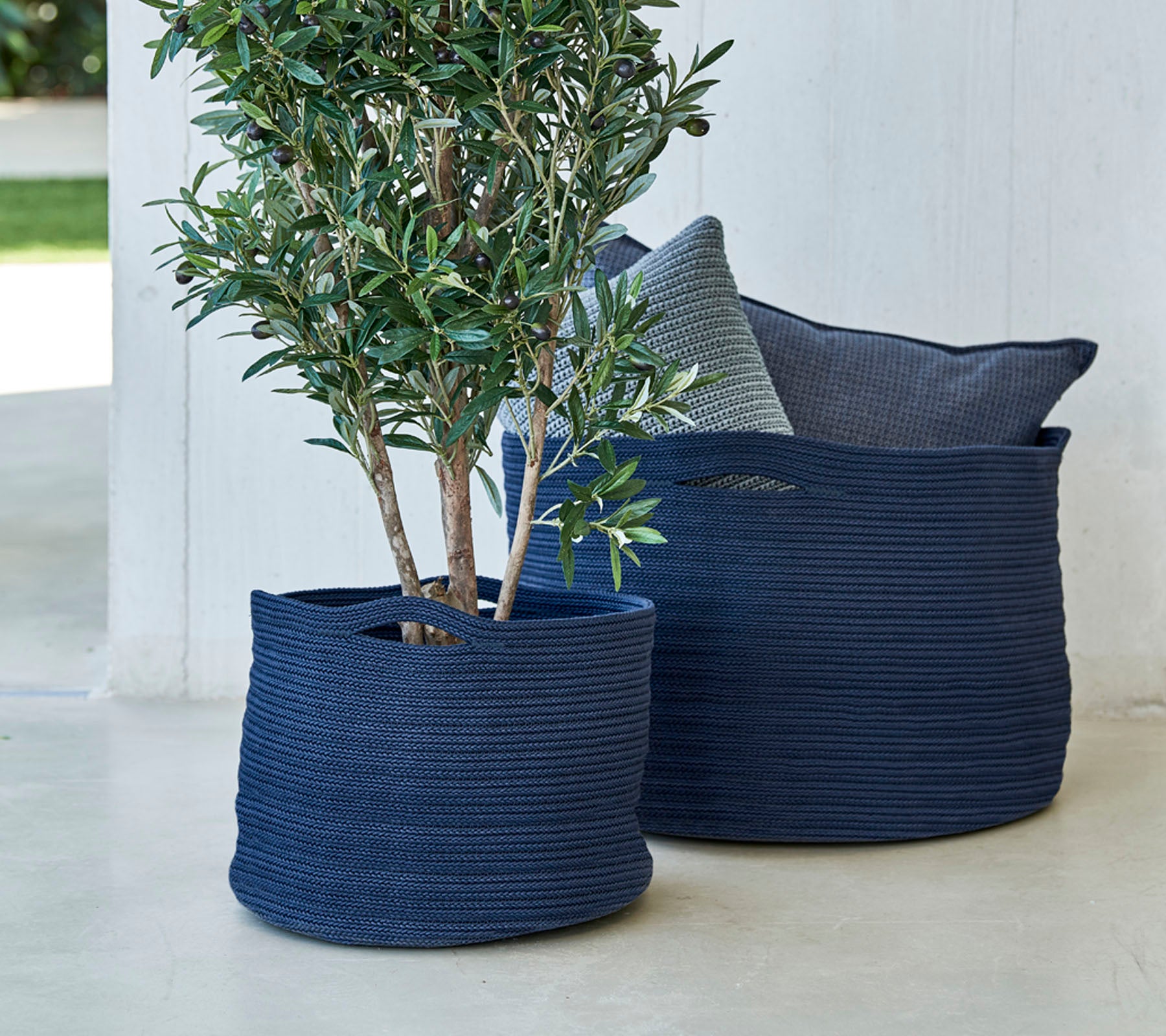 Deux jardinières tissées bleu marine, l'une avec un petit arbre, accompagnées d'un coussin décoratif.