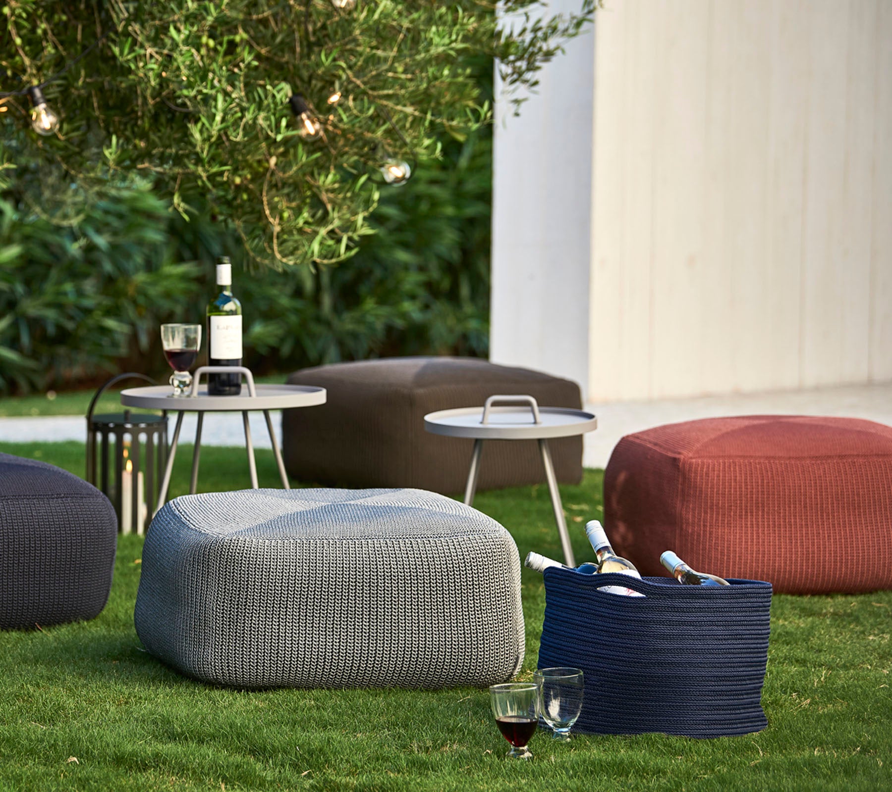 Des poufs texturés de différentes couleurs disposés sur l'herbe, accompagnés d'une petite table avec des boissons et des bouteilles de vin.