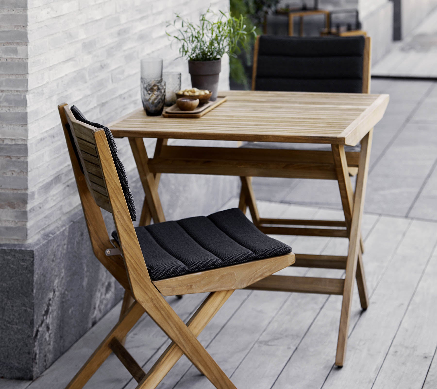 Ensemble table et chaises en bois avec coussins noirs, agrémenté de plantes en pot et de verreries décoratives. Parfait pour une ambiance cosy.