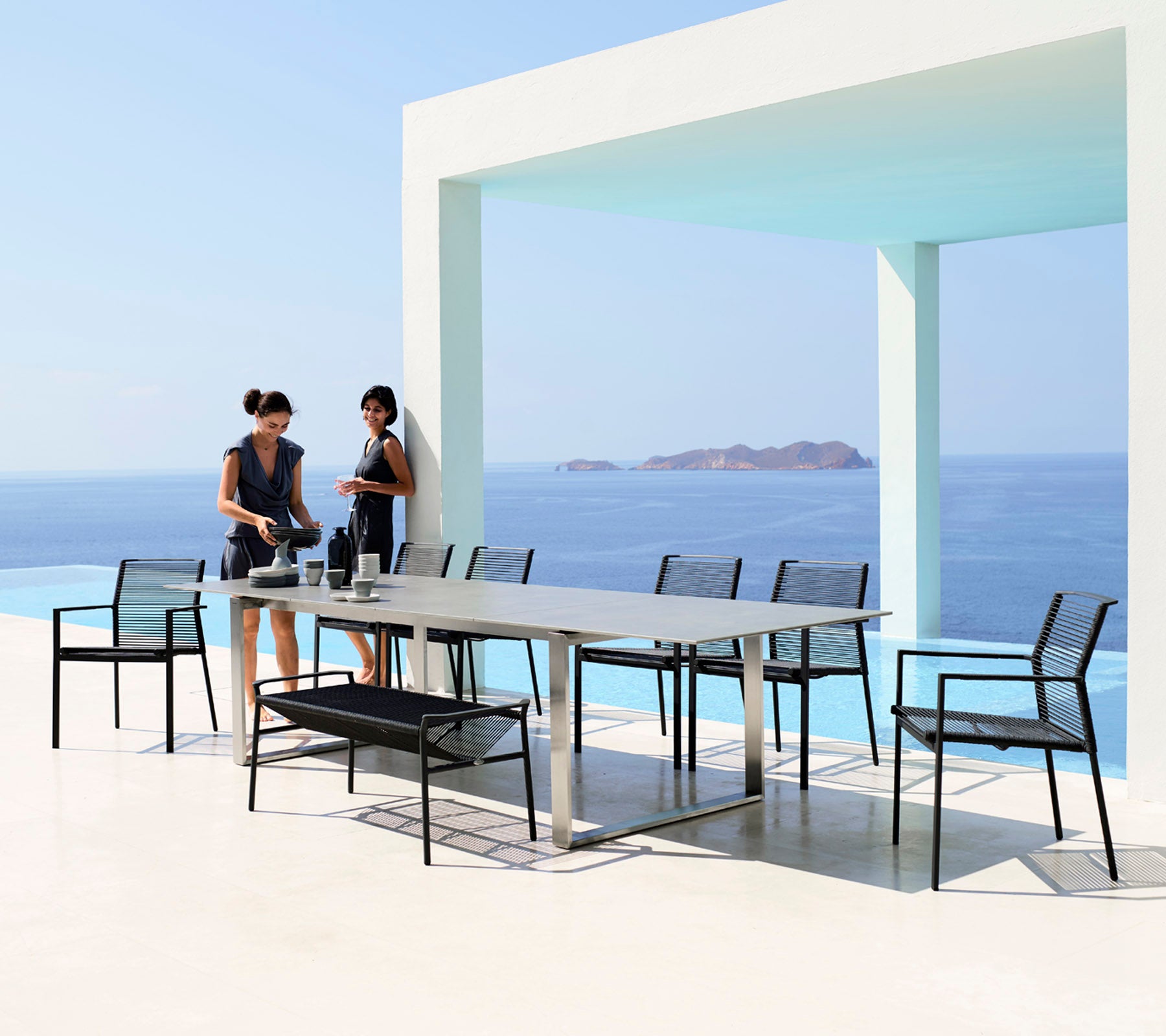Deux femmes préparent une table à manger avec de élégantes chaises noires sur fond d'une mer et d'un ciel bleus sereins.