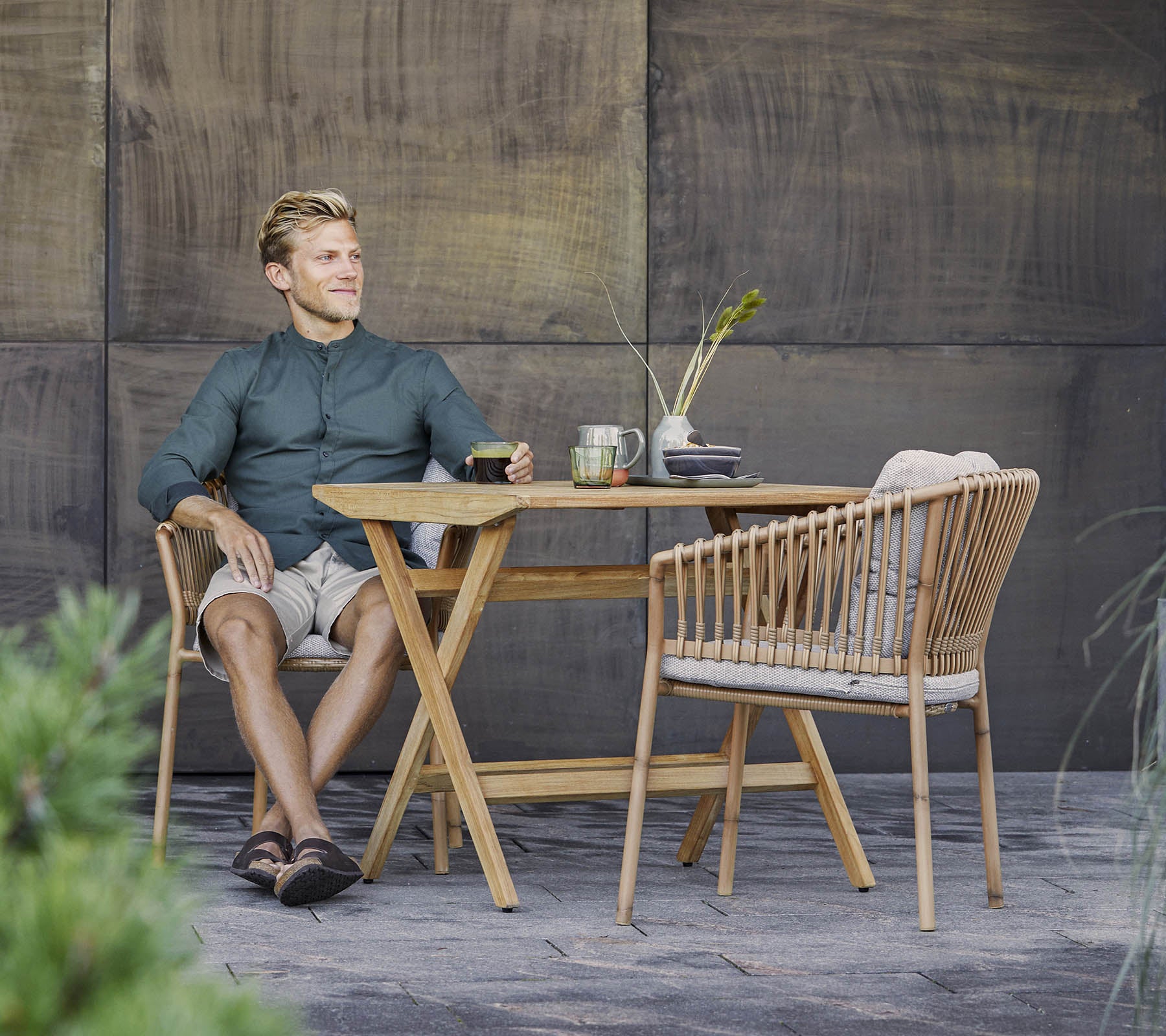 Un homme est assis détendu à une table en bois élégante avec une boisson légère, entouré de verdure et d'un fond texturé.