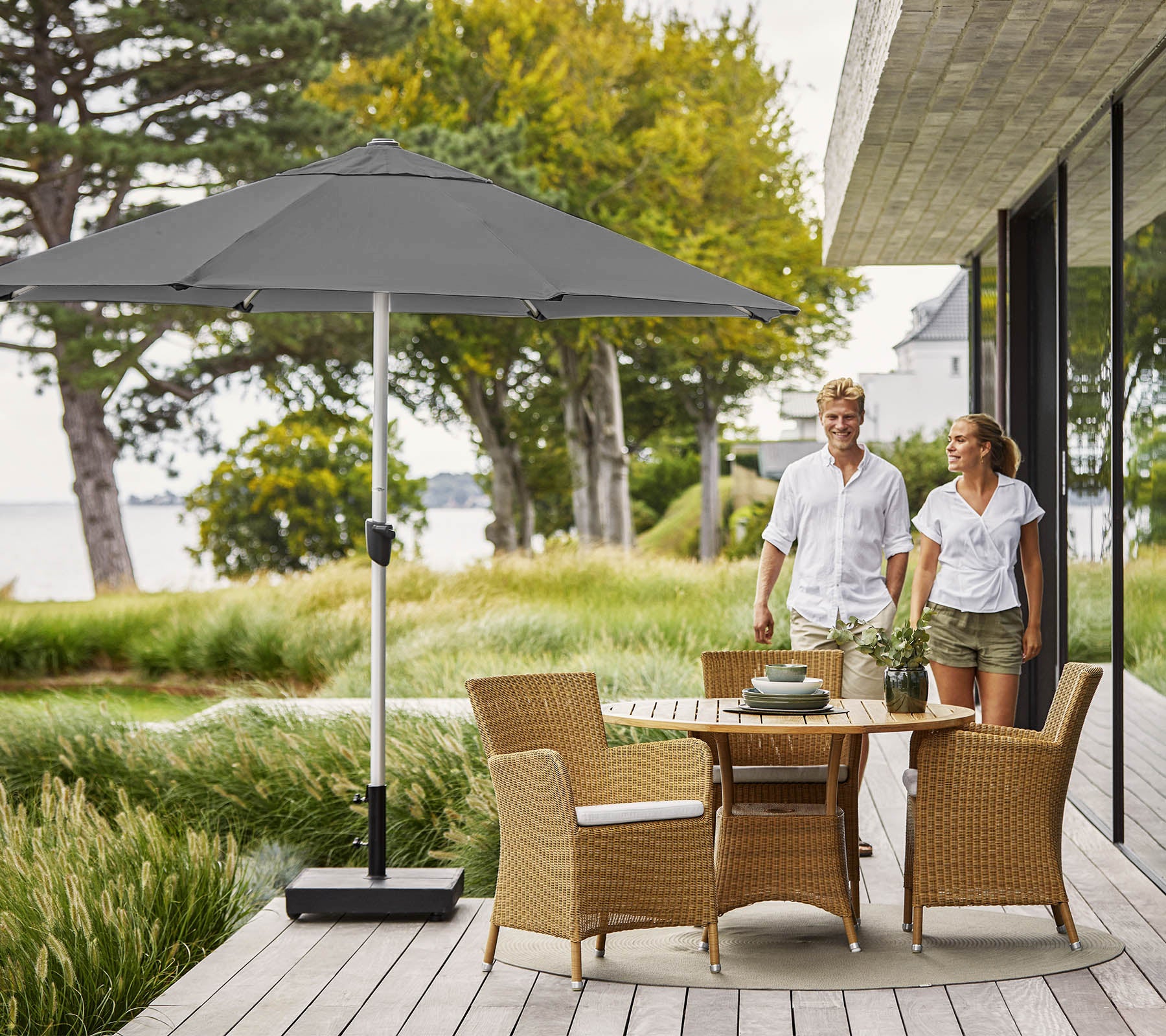 Un couple marchant sur une terrasse en bois avec une table ronde et des chaises en osier, ombragé par un grand parasol gris, surplombant une verdure luxuriante.