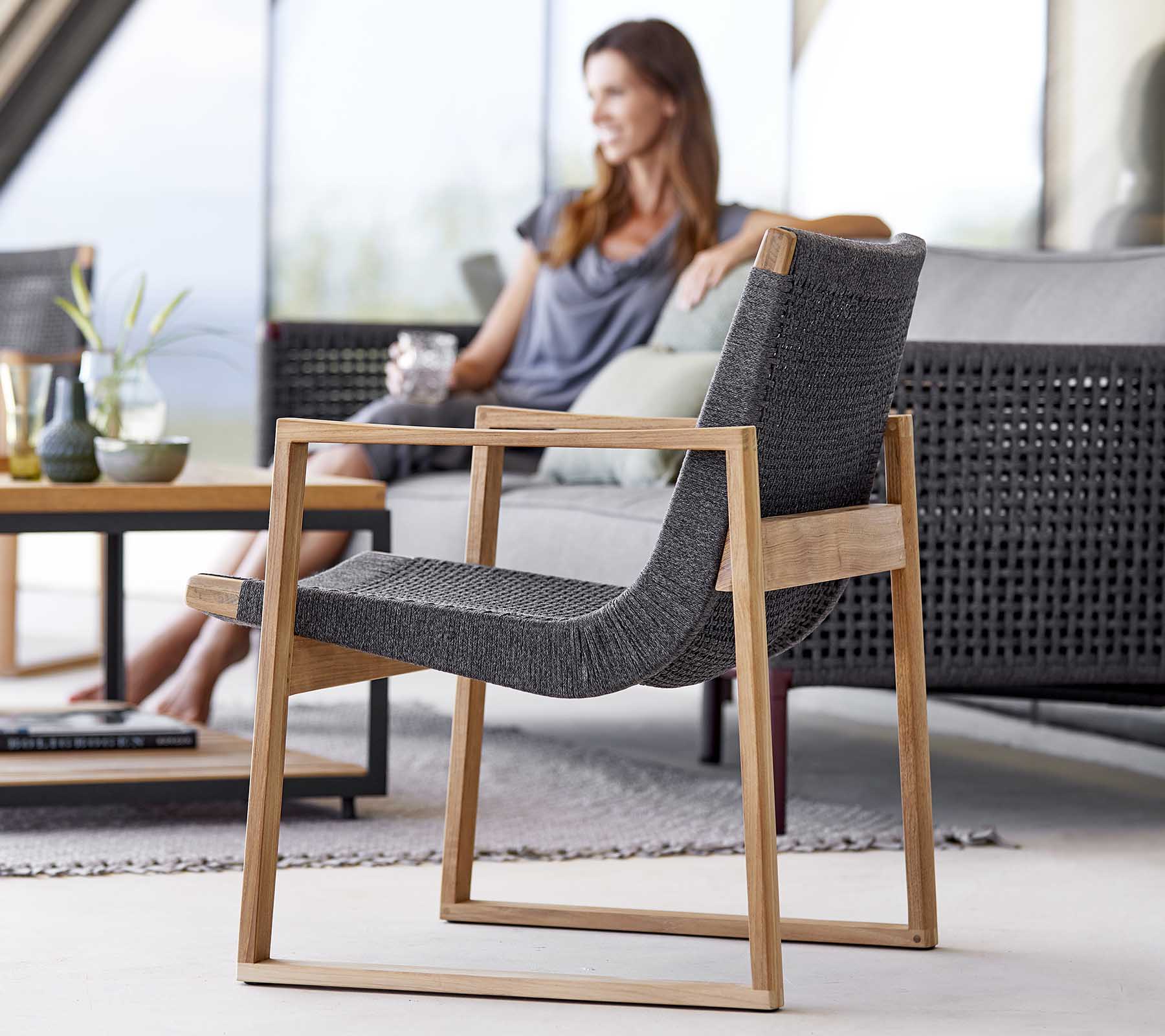 Une chaise moderne avec un cadre en bois lisse et un tissu tissé, positionnée à côté d'une femme qui se détend et apprécie une boisson.