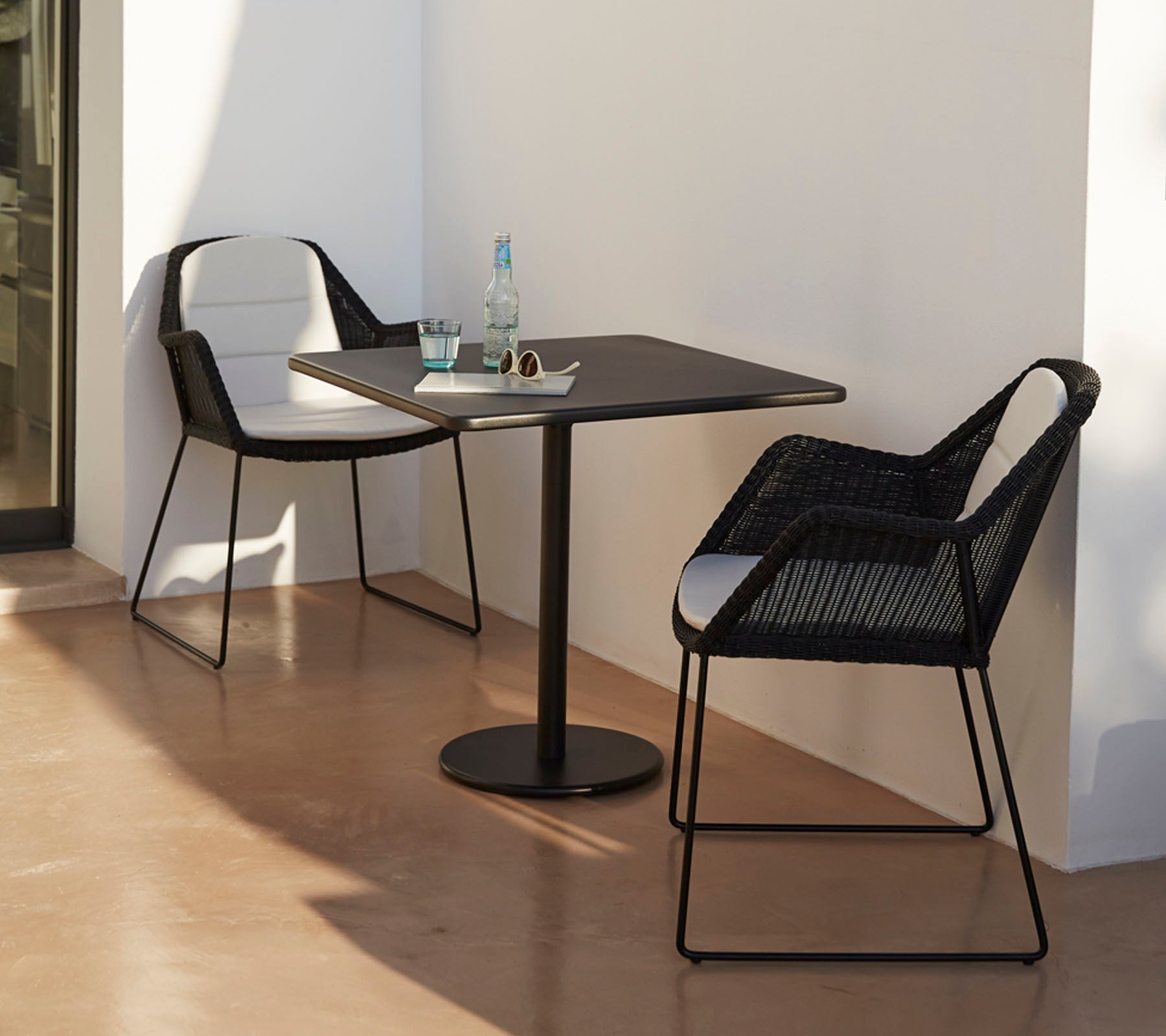 Table moderne avec deux élégantes chaises noires, accompagnée d'une bouteille d'eau et de lunettes de soleil, placée sur un fond minimaliste.