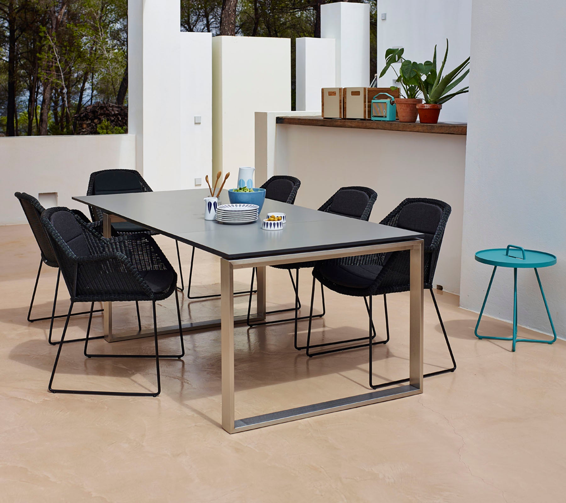 Un agencement de salle à manger moderne avec une table rectangulaire à la surface lisse, entourée de six chaises tressées noires et d'une petite table d'appoint teal.
