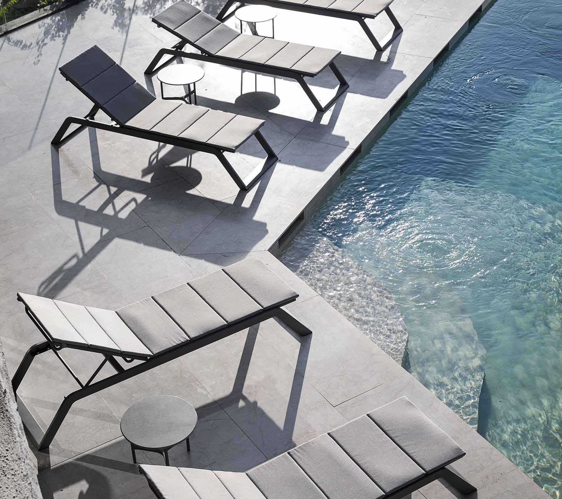 Une rangée de chaises longues modernes avec une petite table d'appoint à côté d'une piscine, projetant des ombres sur le pavé.
