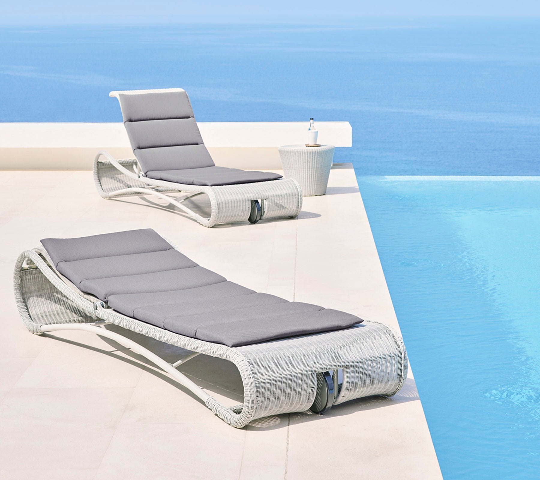 Deux fauteuils lounge élégants avec des coussins gris à côté d'une paisible piscine à débordement bleue, créant un espace extérieur relaxant.