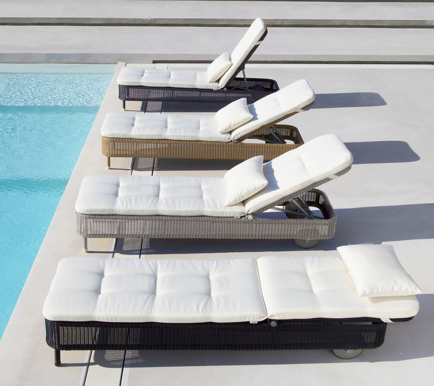Quatre chaises longues de différentes couleurs avec des coussins blancs, placées à côté d'une piscine, chacune avec un dossier réglable.