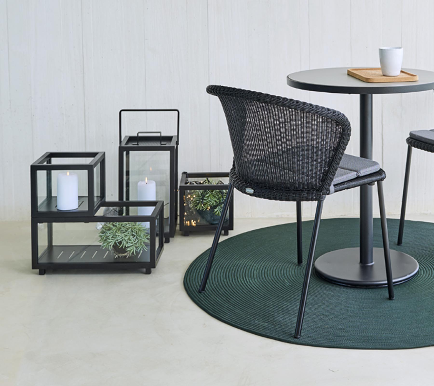 Des sièges modernes et des lanternes en verre décoratives avec des plantes, disposées autour d'une table ronde, sur un tapis rond vert.