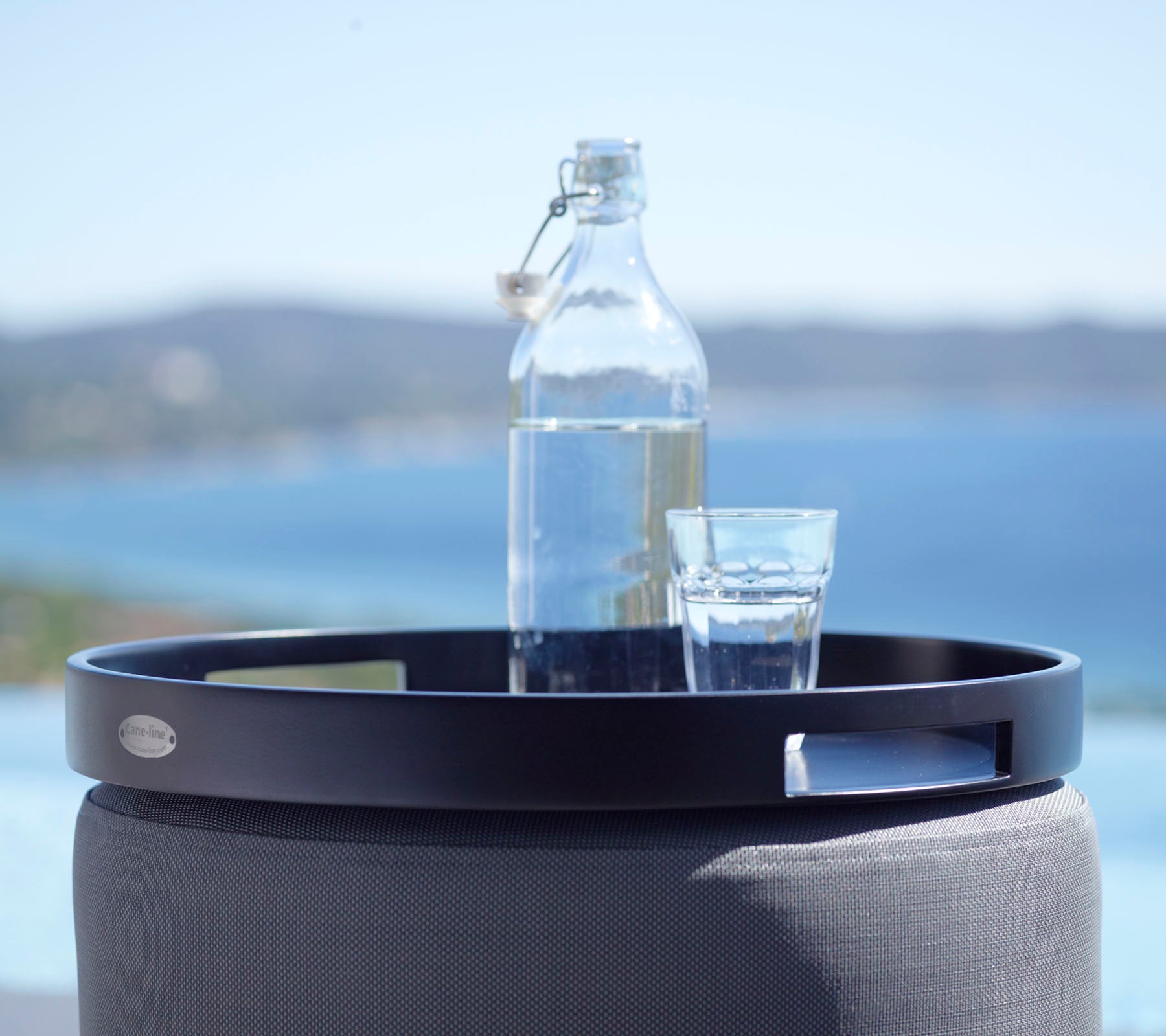 Un plateau rond noir tient un verre d’eau et une bouteille en verre, sur fond de mer et de collines lointaines.
