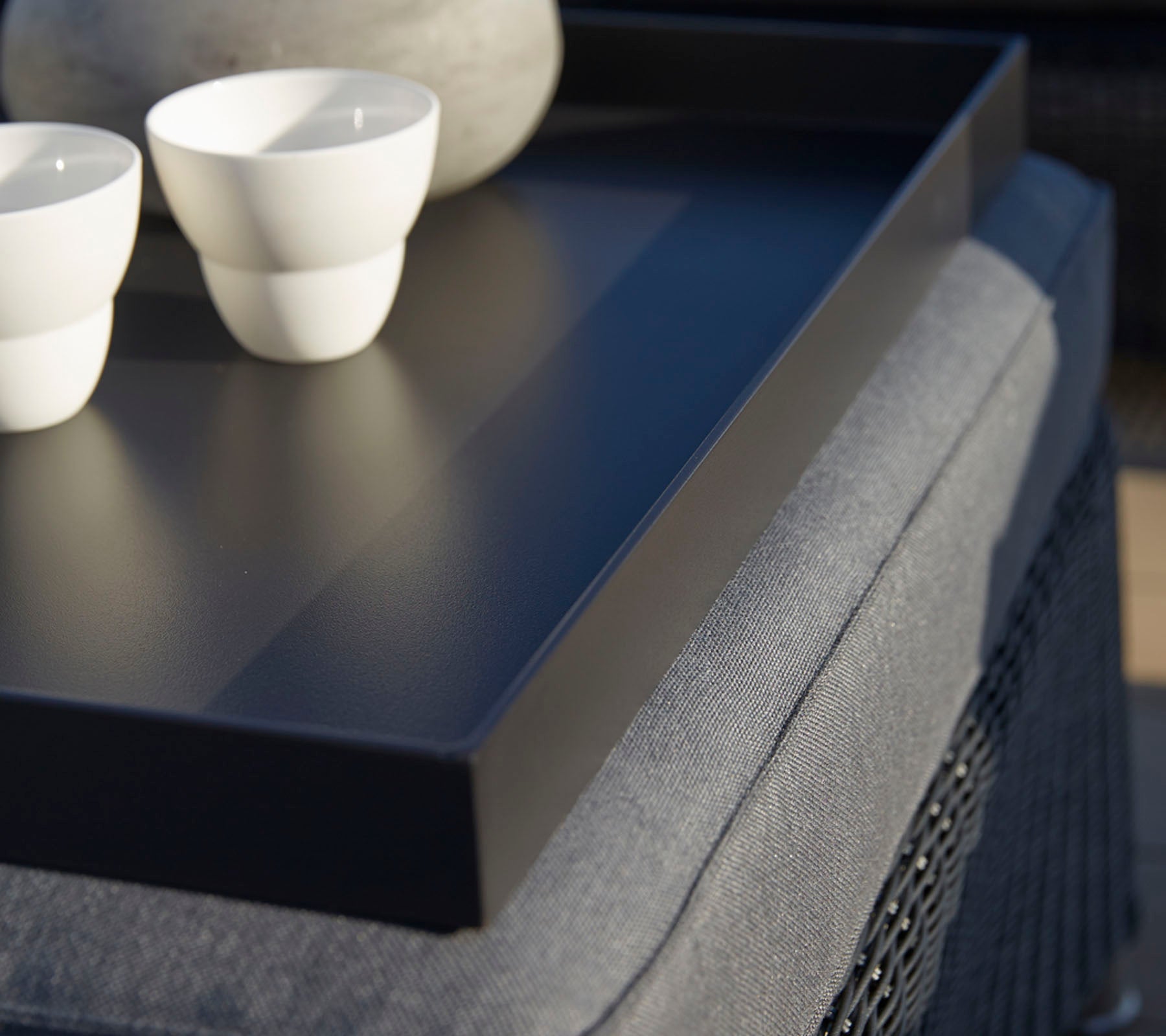 Un plateau noir contient deux tasses blanches et une pierre lisse, positionnées sur une surface texturée. Un éclairage doux renforce la scène.