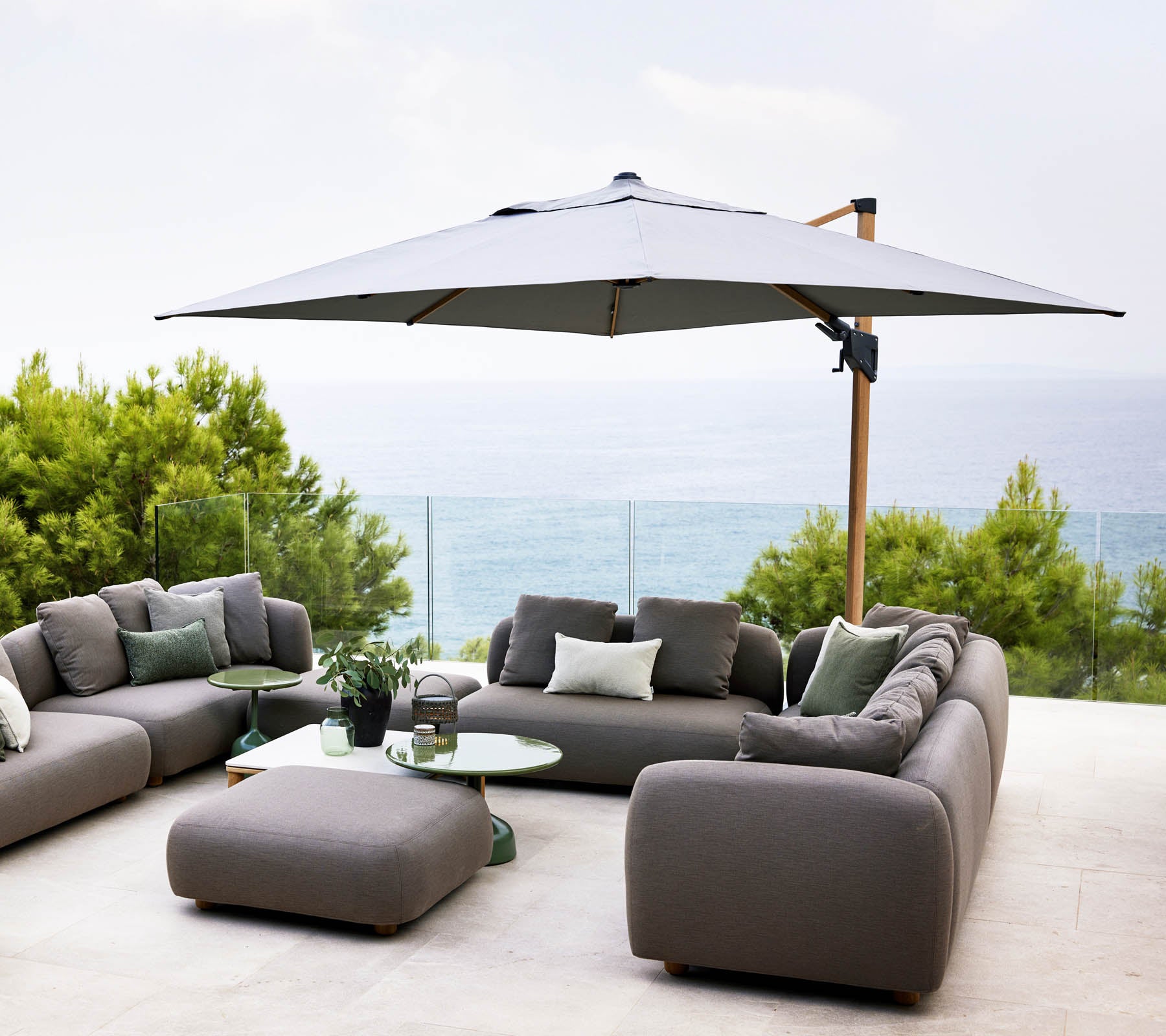Espace extérieur élégant avec un grand parasol, des canapés gris et une table en verre, surplombant une vue sereine sur l'eau et une verdure luxuriante.