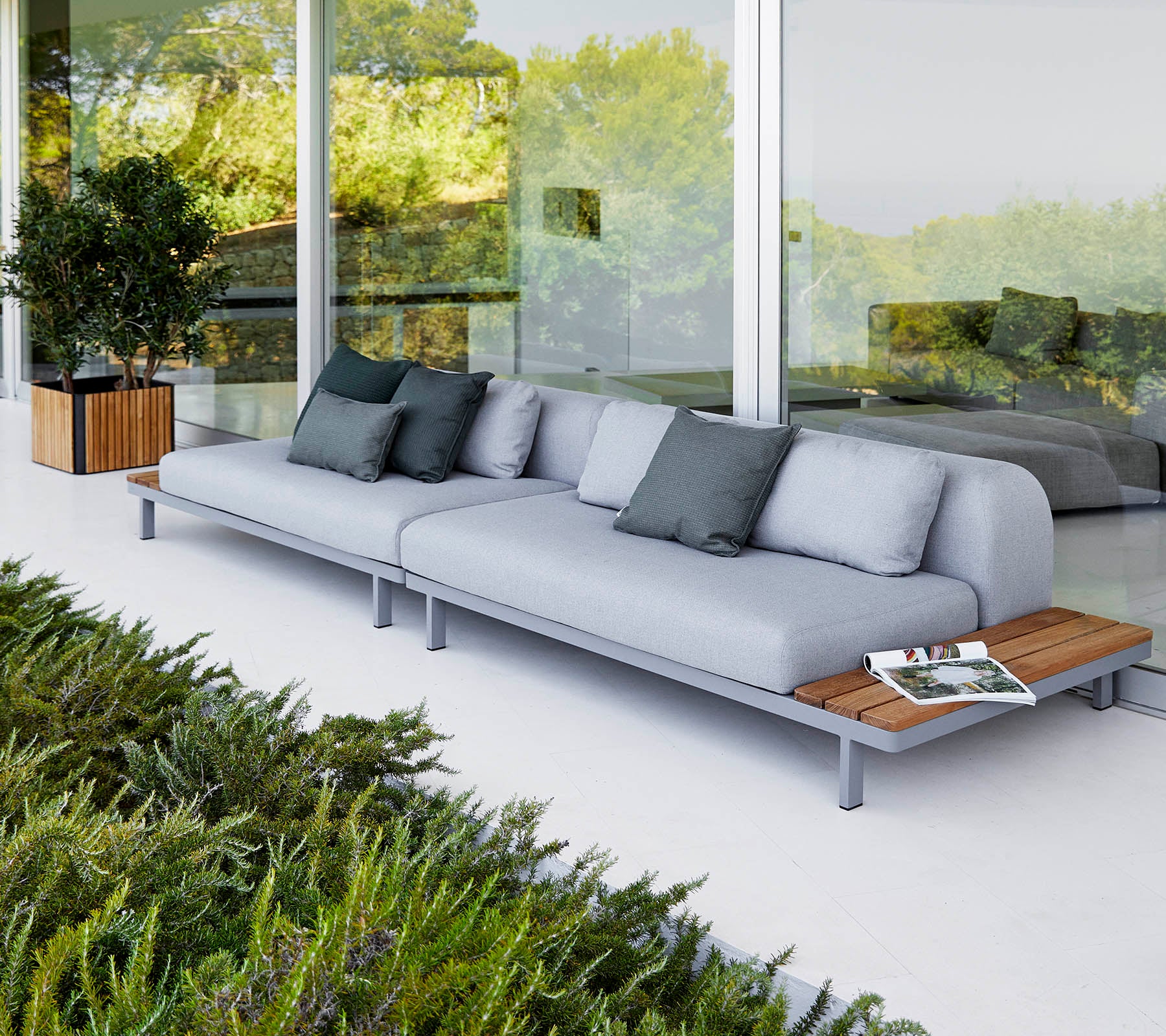 Sofa moderne avec des coussins gris clair et des accents en bois, accompagnée de coussins décoratifs et entourée de verdure.