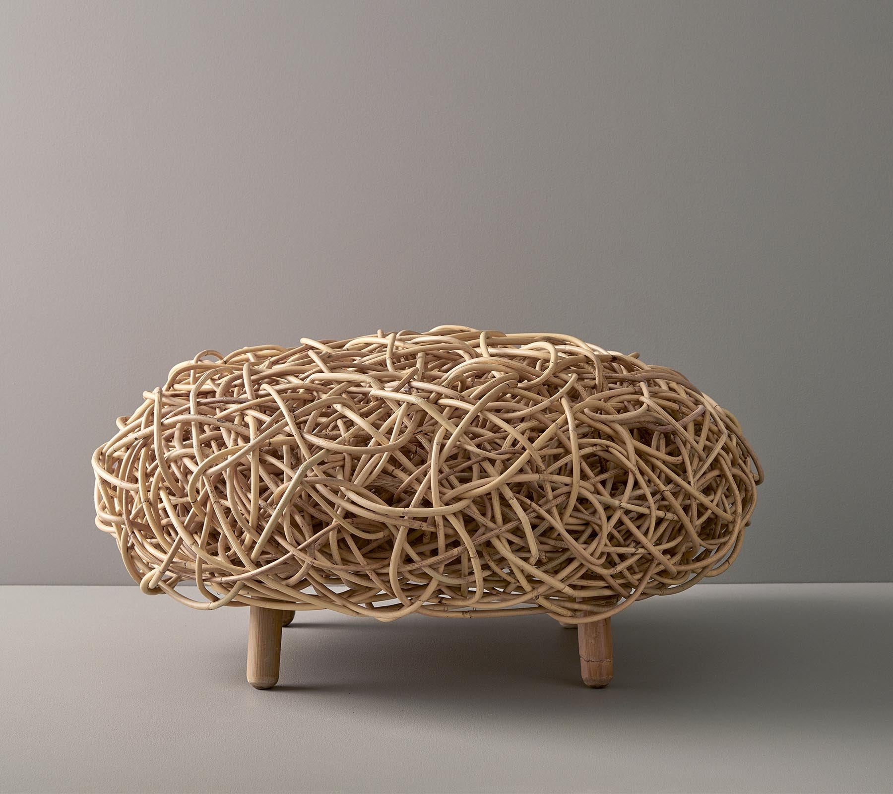 Pièce décorative tissée naturellement avec une forme ovale, présentant des brins entrelacés complexes et soutenue par de courtes pattes en bois.