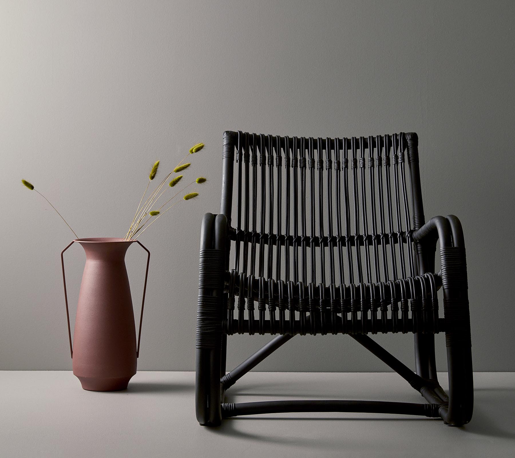 Chaise tissée noire élégante à côté d'un vase rose avec des tiges vertes, sur fond gris minimaliste.