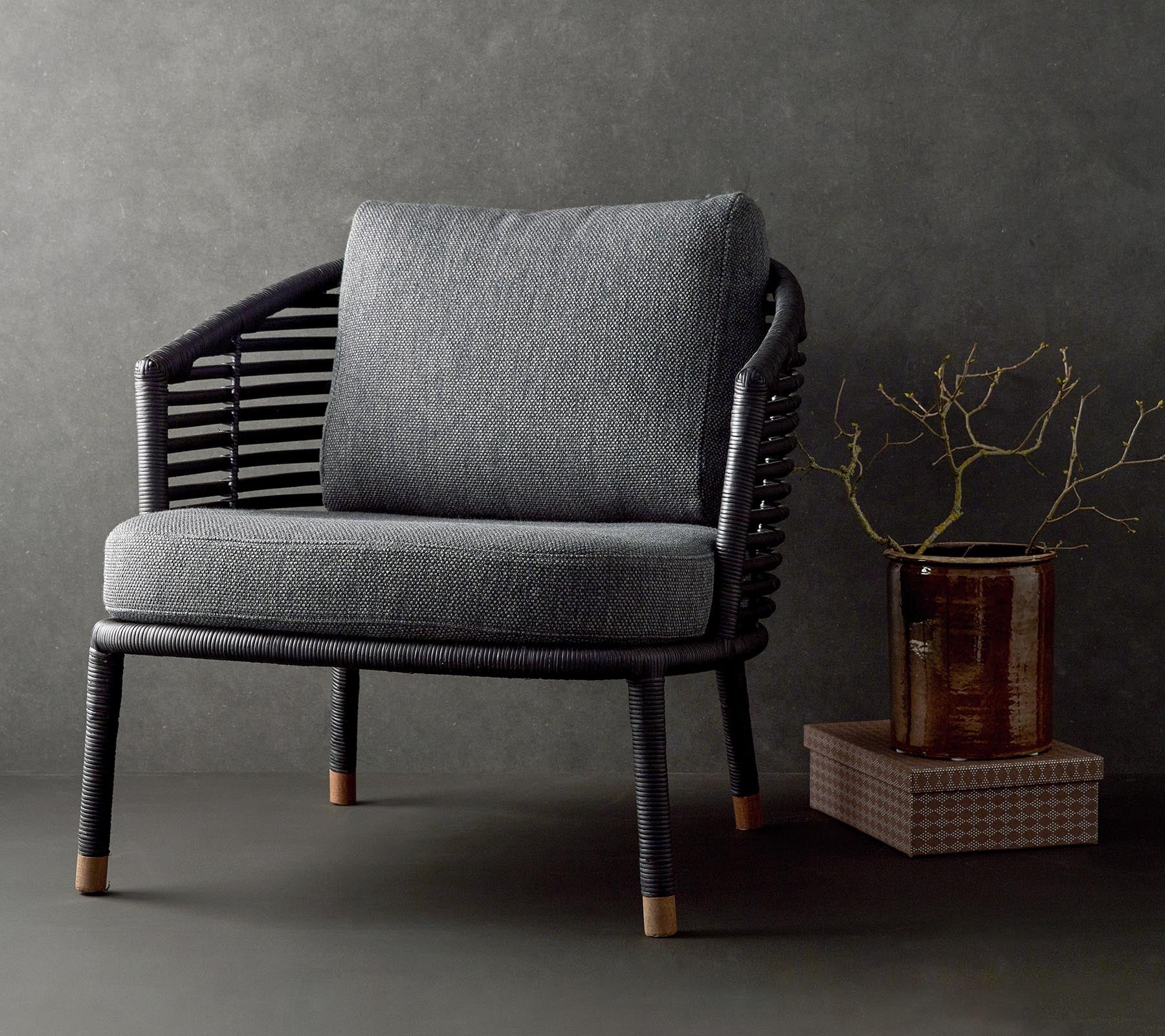 Chaise élégante avec un coussin gris texturé et un cadre noir tissé, complétée par une branche décorative et une petite table.