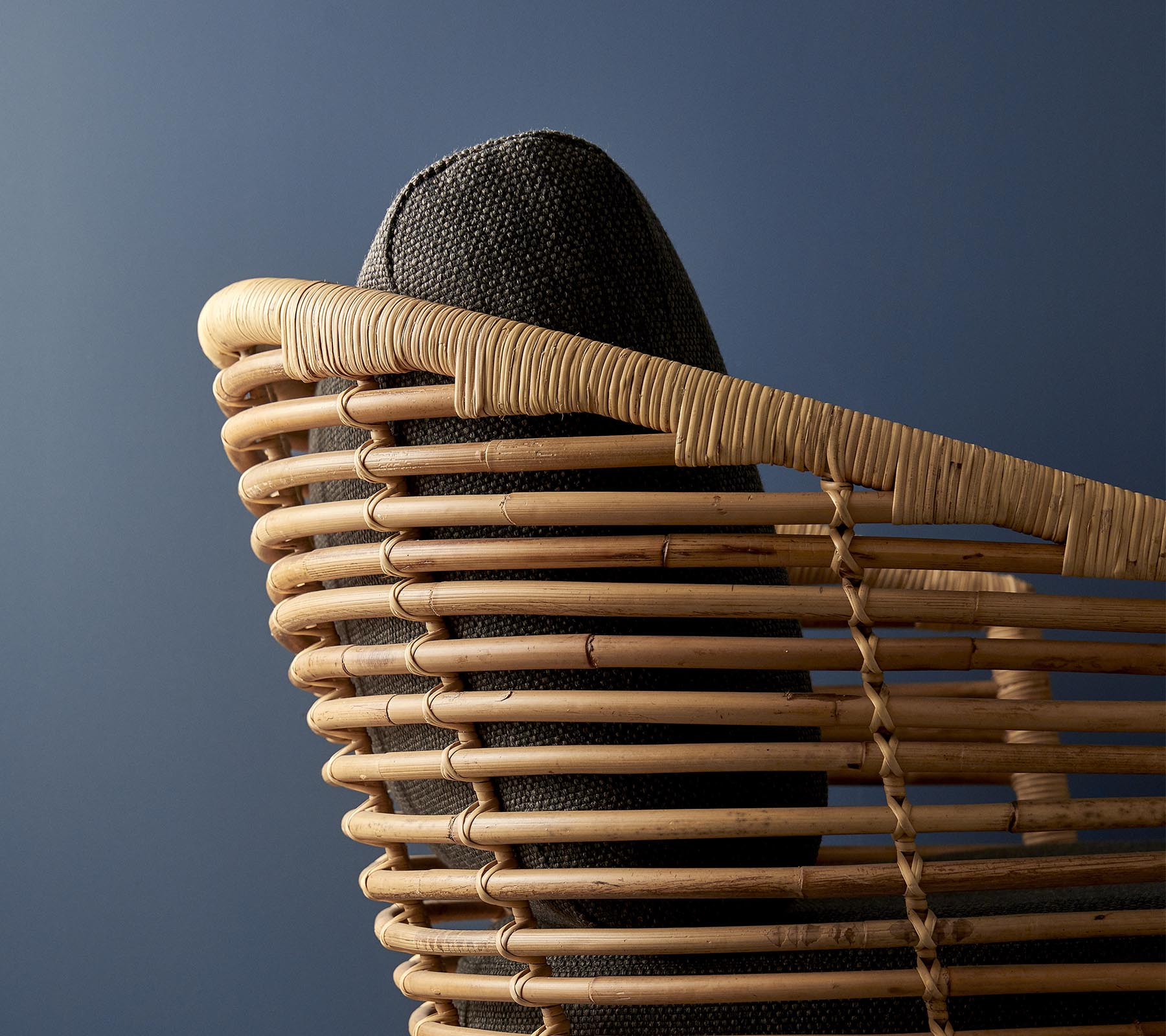 Chaise tissée élégante avec un dossier incurvé et un siège rembourré foncé sur un fond bleu atténué.