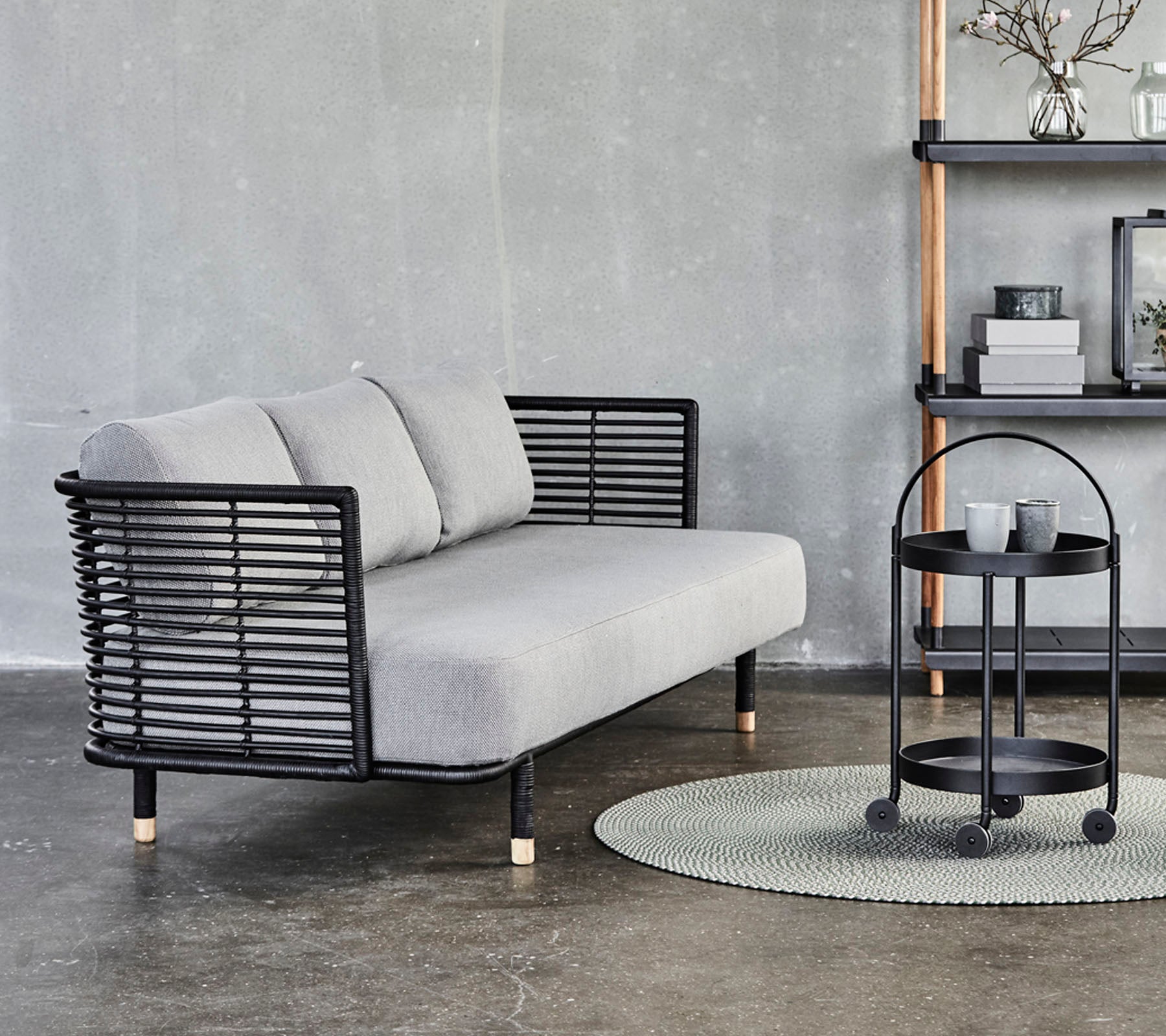 Canapé gris élégant avec des accents en métal noir et des coussins, associé à une table d'appoint ronde sur un tapis texturé contre un fond neutre.