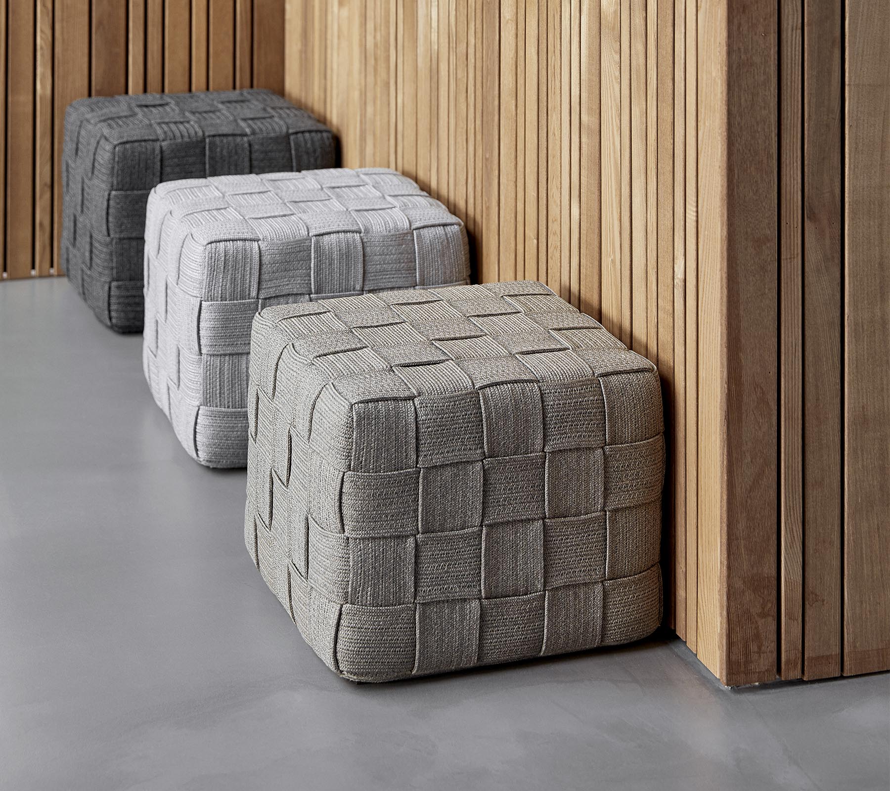 Trois ottomans carrés texturés dans des nuances de gris variées sont positionnés contre un mur en bois, ajoutant une touche moderne à l'ensemble.