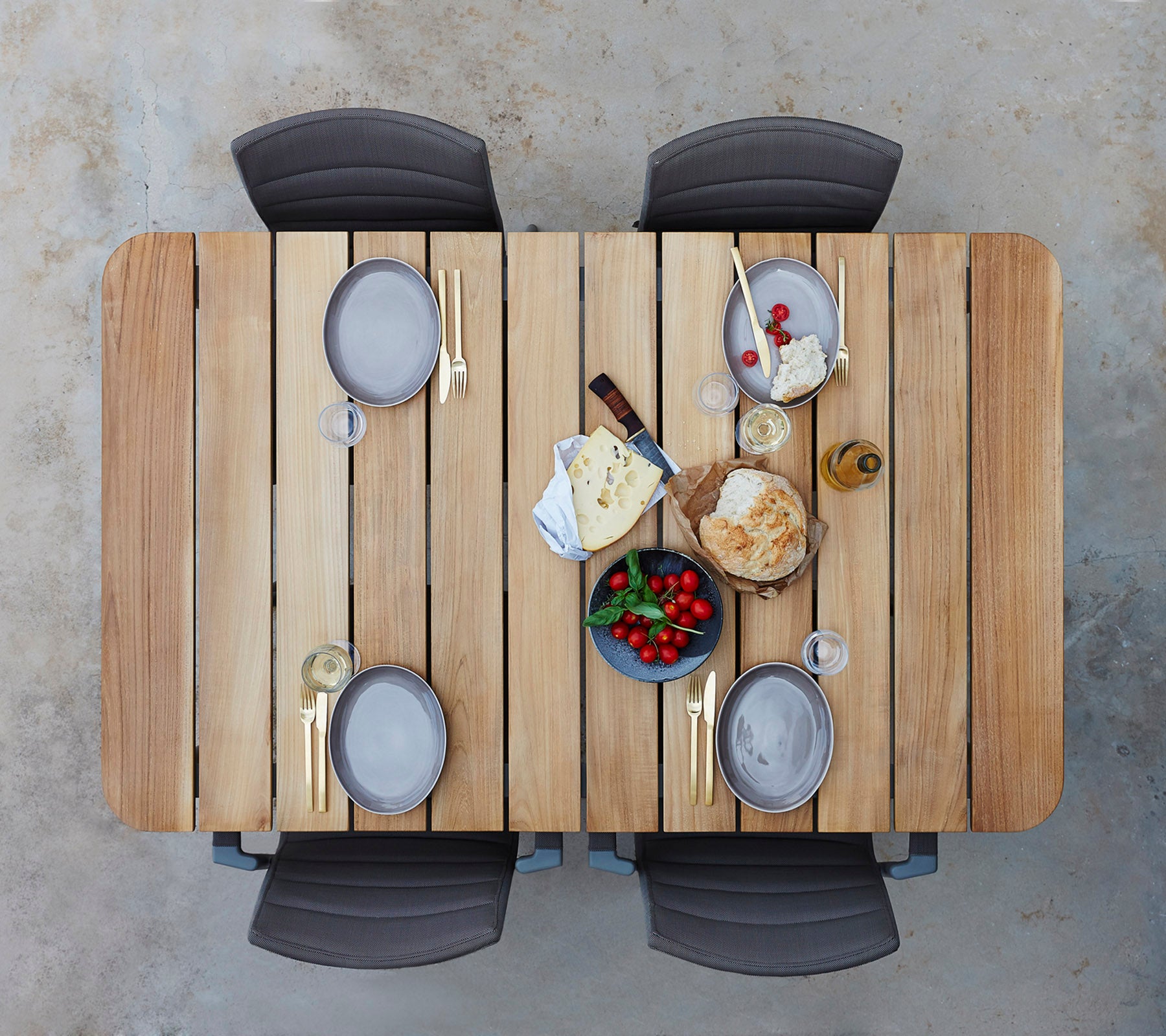 Une table en bois dressée avec des assiettes, des verres et un assortiment de fromages, de fruits et de pain, entourée de quatre chaises.