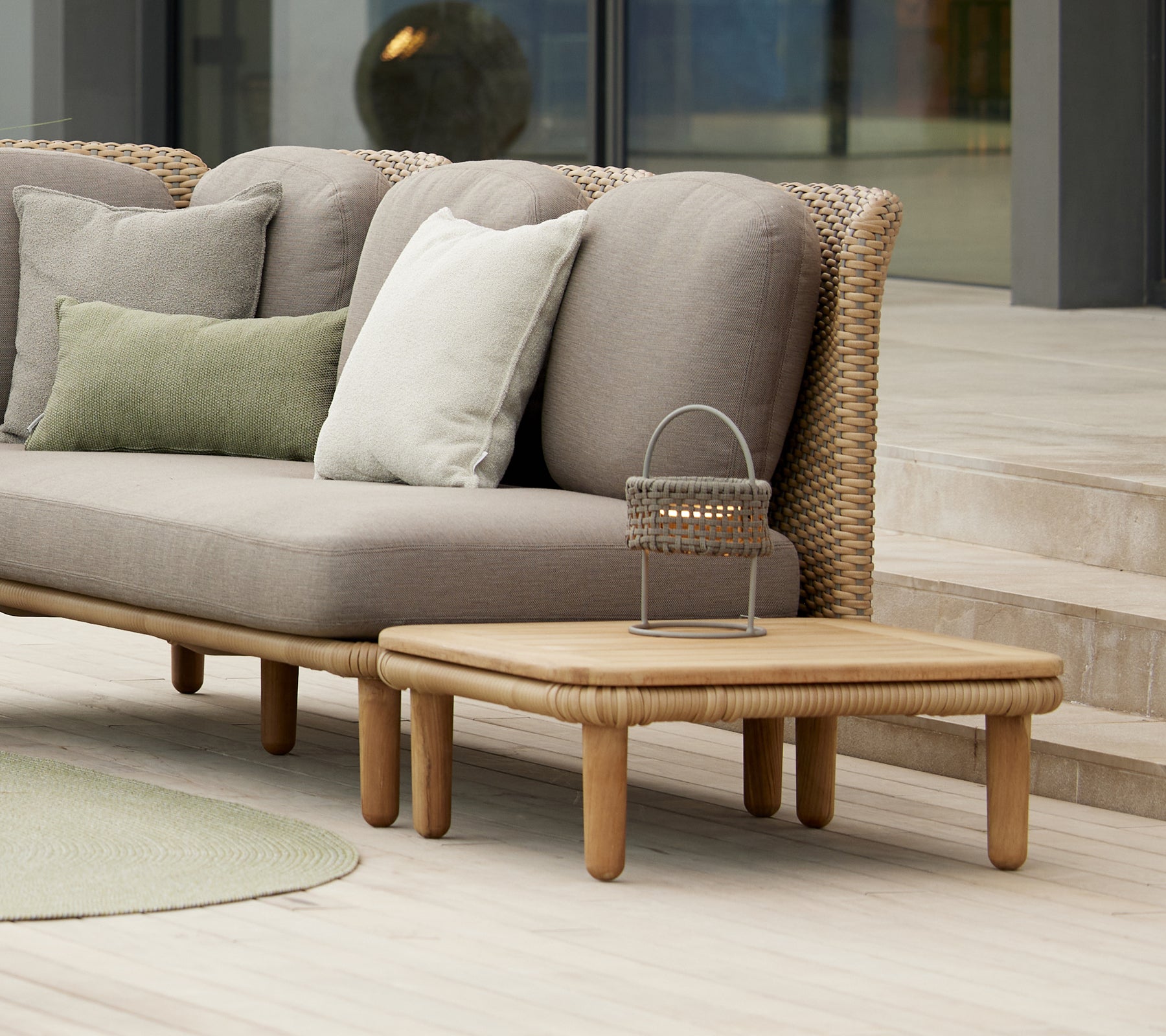 Table basse élégante en arc avec un plateau en teck à côté de coussins sur un canapé moderne, complétée par une lampe décorative Illusion.