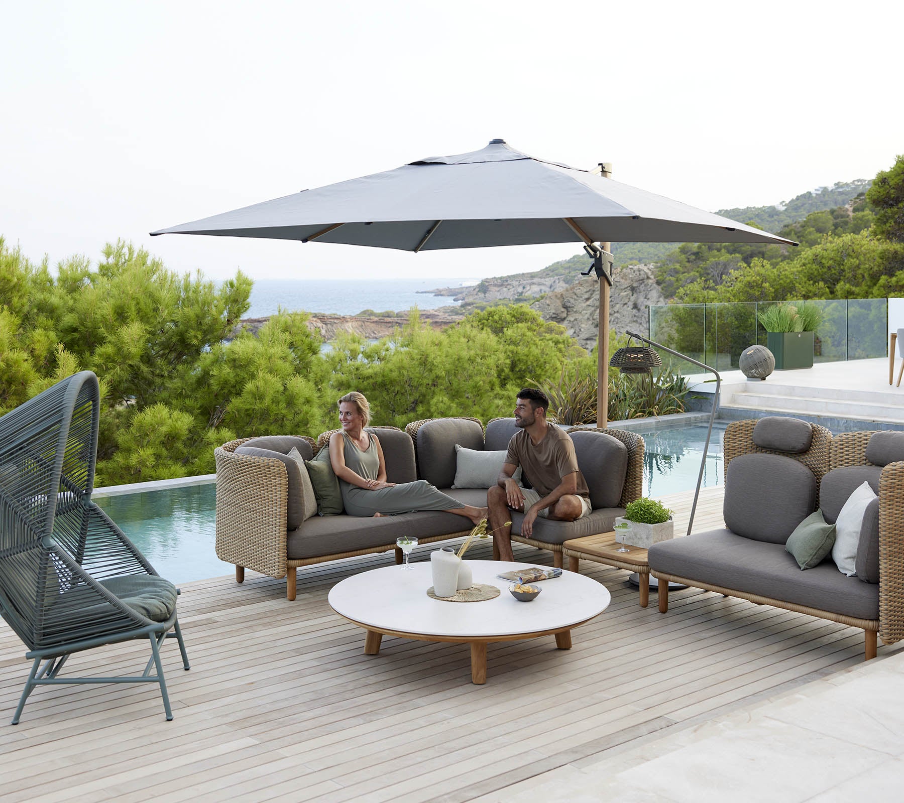 Un salon élégant avec des sièges confortables, un grand parasol et une table ronde, entouré de verdure et d'une vue sereine.