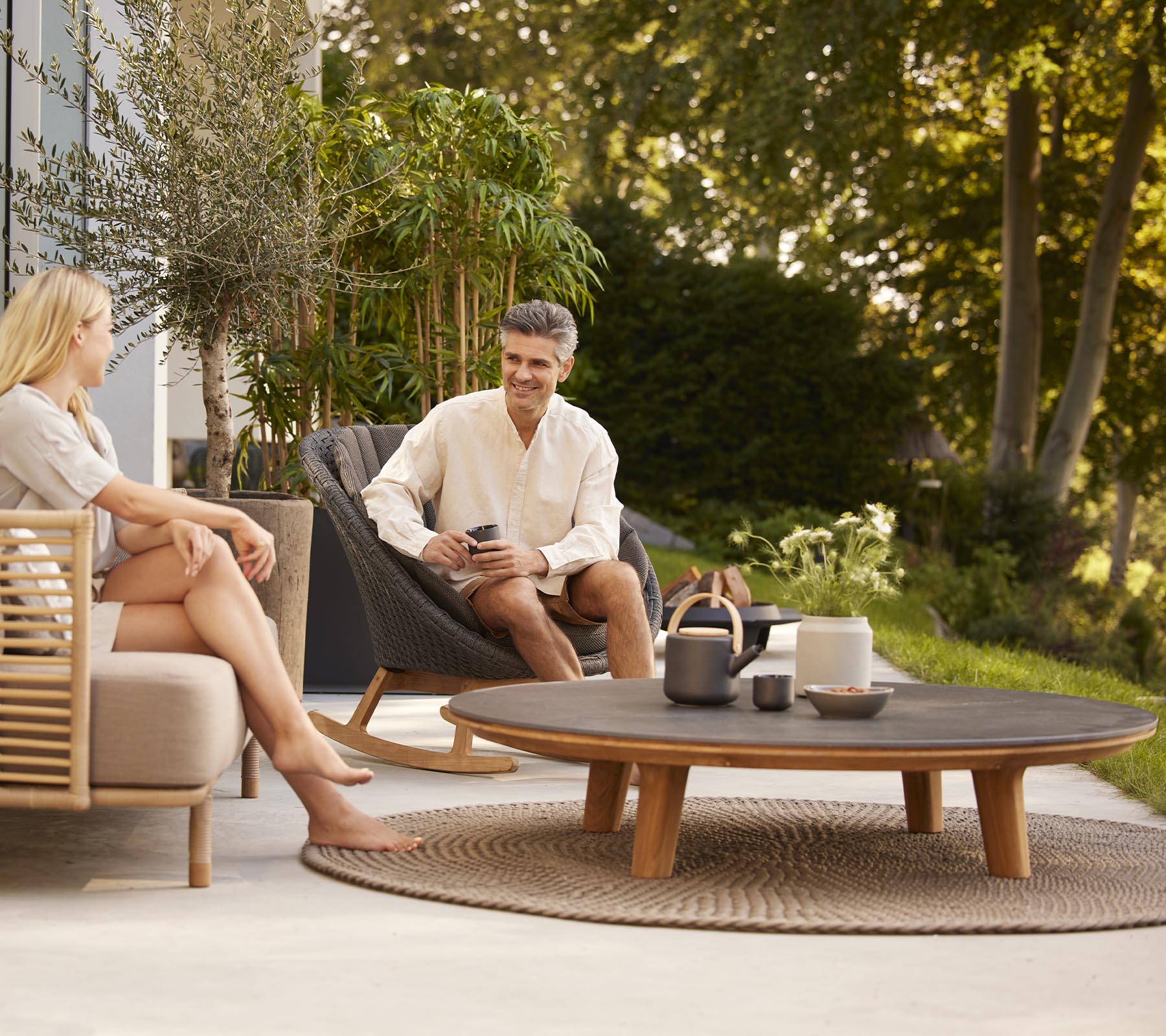 Un homme et une femme se détendent sur des sièges confortables, appréciant des boissons sur une table basse ronde entourée de verdure.