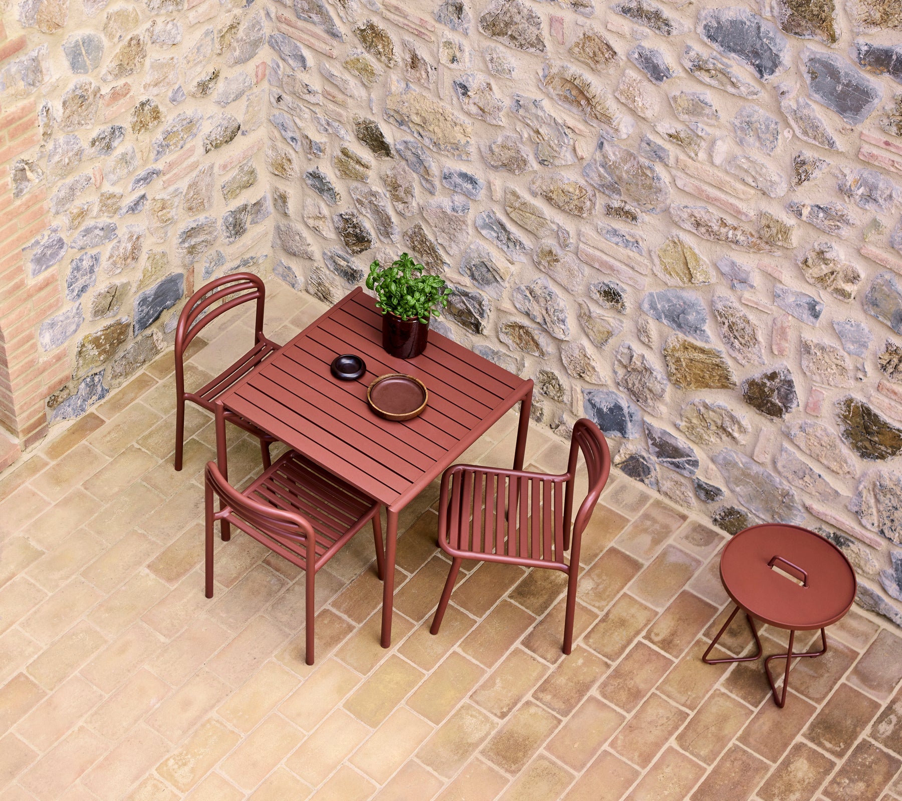 Une table et des chaises dans une teinte rouge désert, à côté d'une petite table ronde, contre un mur en pierre texturé avec une plante en pot.