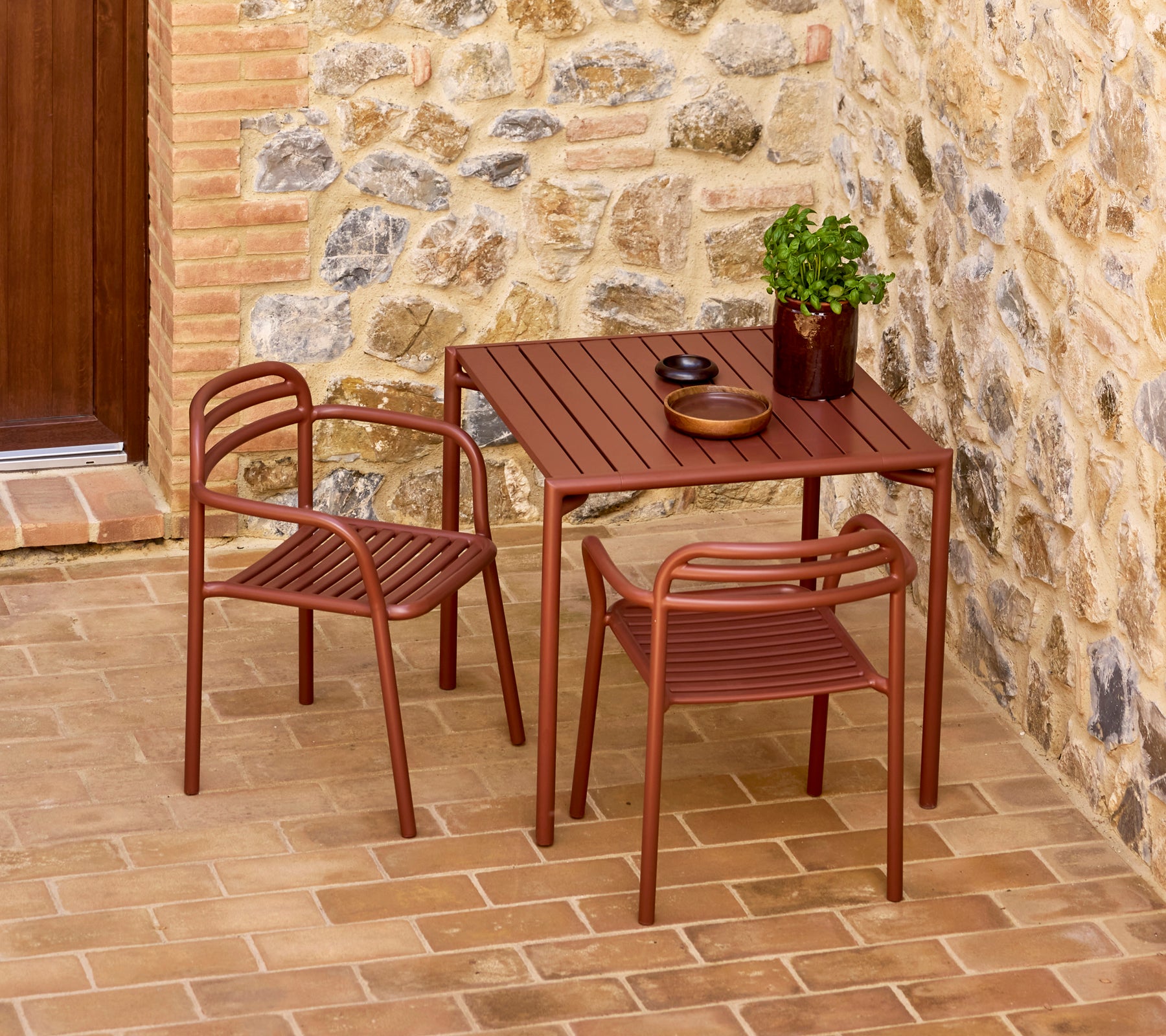 Table Bliss rouge désert élégante avec deux chaises assorties, accompagnée d'une plante décorative et de vaisselle simple sur un fond en pierre.