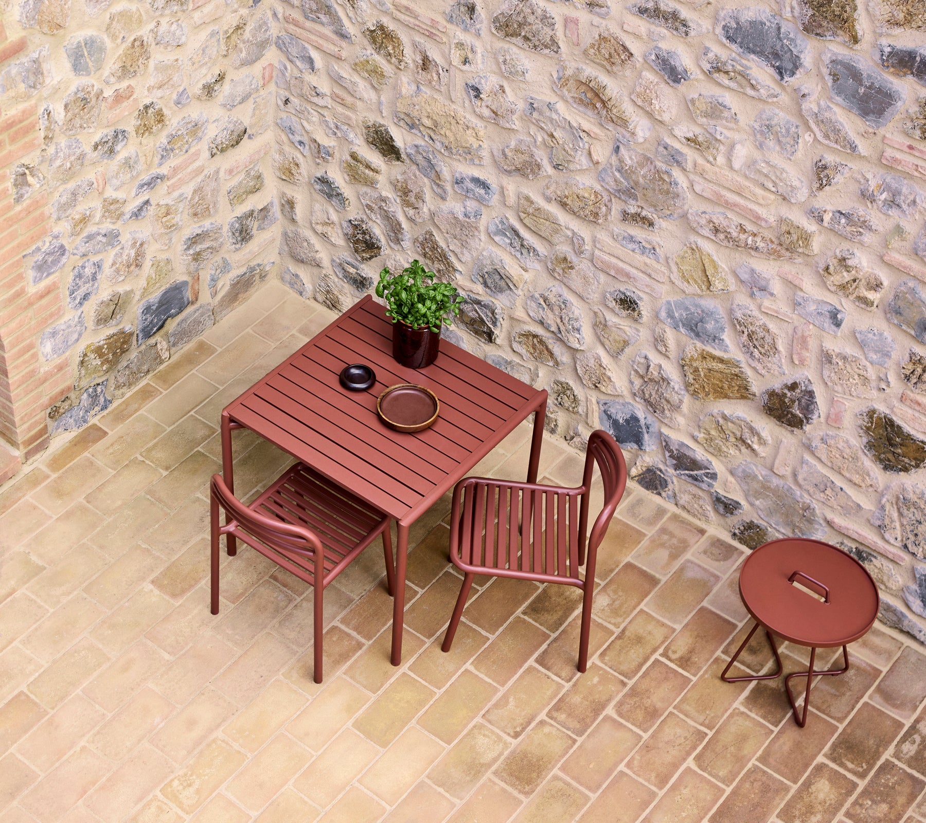 une table de café rouge avec deux chaises, comportant une petite plante et une assiette, devant un mur en pierre.