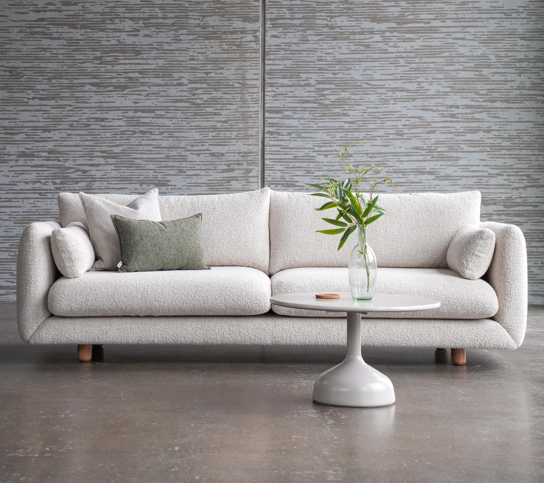 Un canapé élégant avec des coussins à côté d'une table basse Glaxe au dessus rond et jambes en teck élégantes, ornée d'un vase de fleurs.