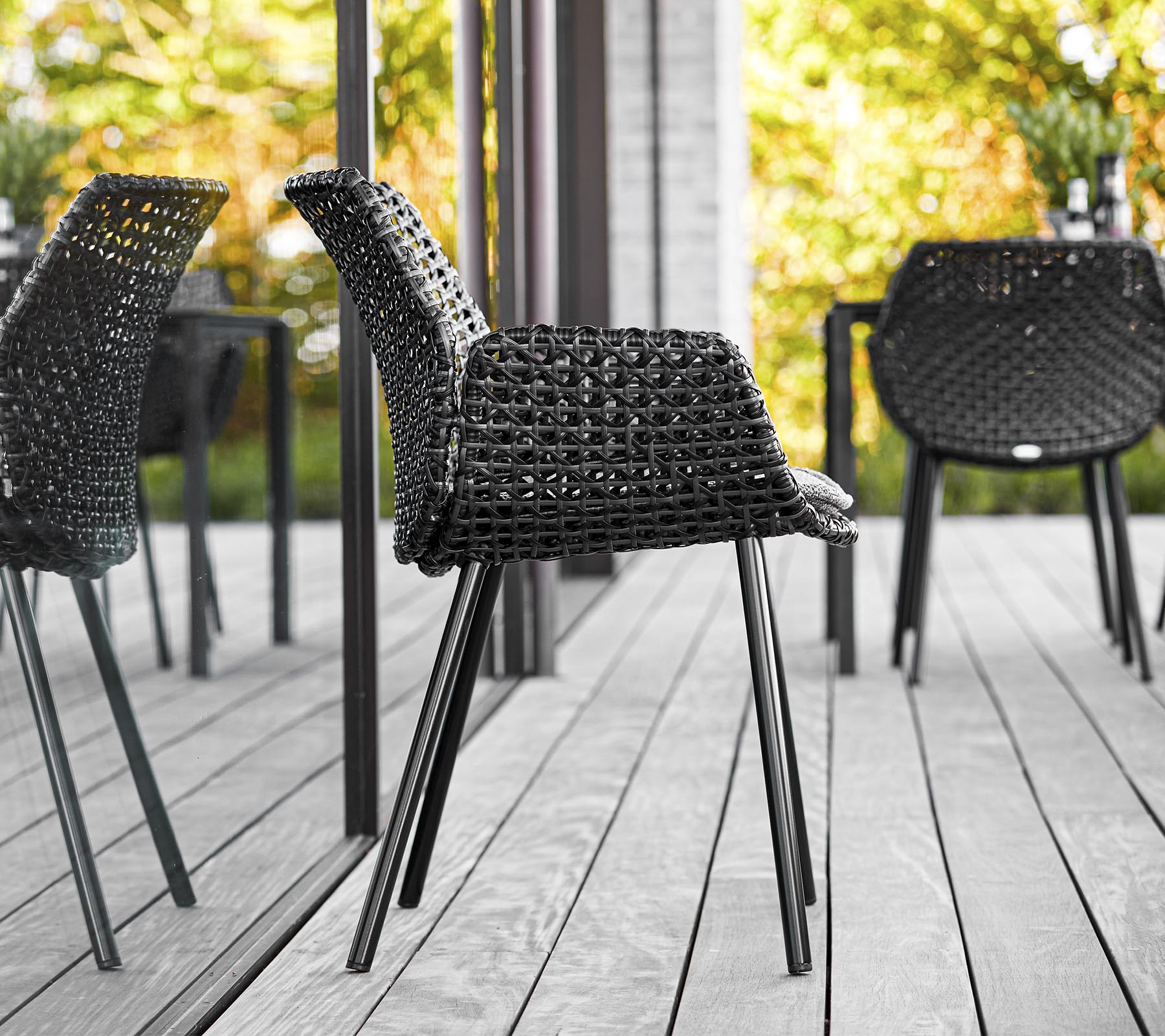 Chaise tressée noire élégante avec des pieds épurés, placée sur un fond de verdure et de reflets, améliorant l'esthétique moderne en extérieur.