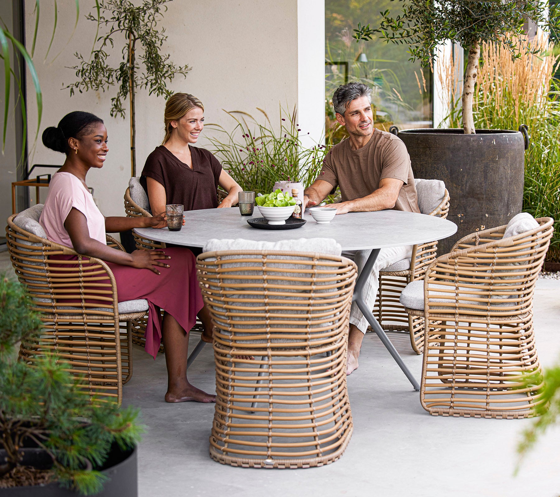 Quatre personnes profitent de boissons autour d'une table ronde moderne, avec des chaises en rattan élégantes et de la verdure dans un cadre serein.