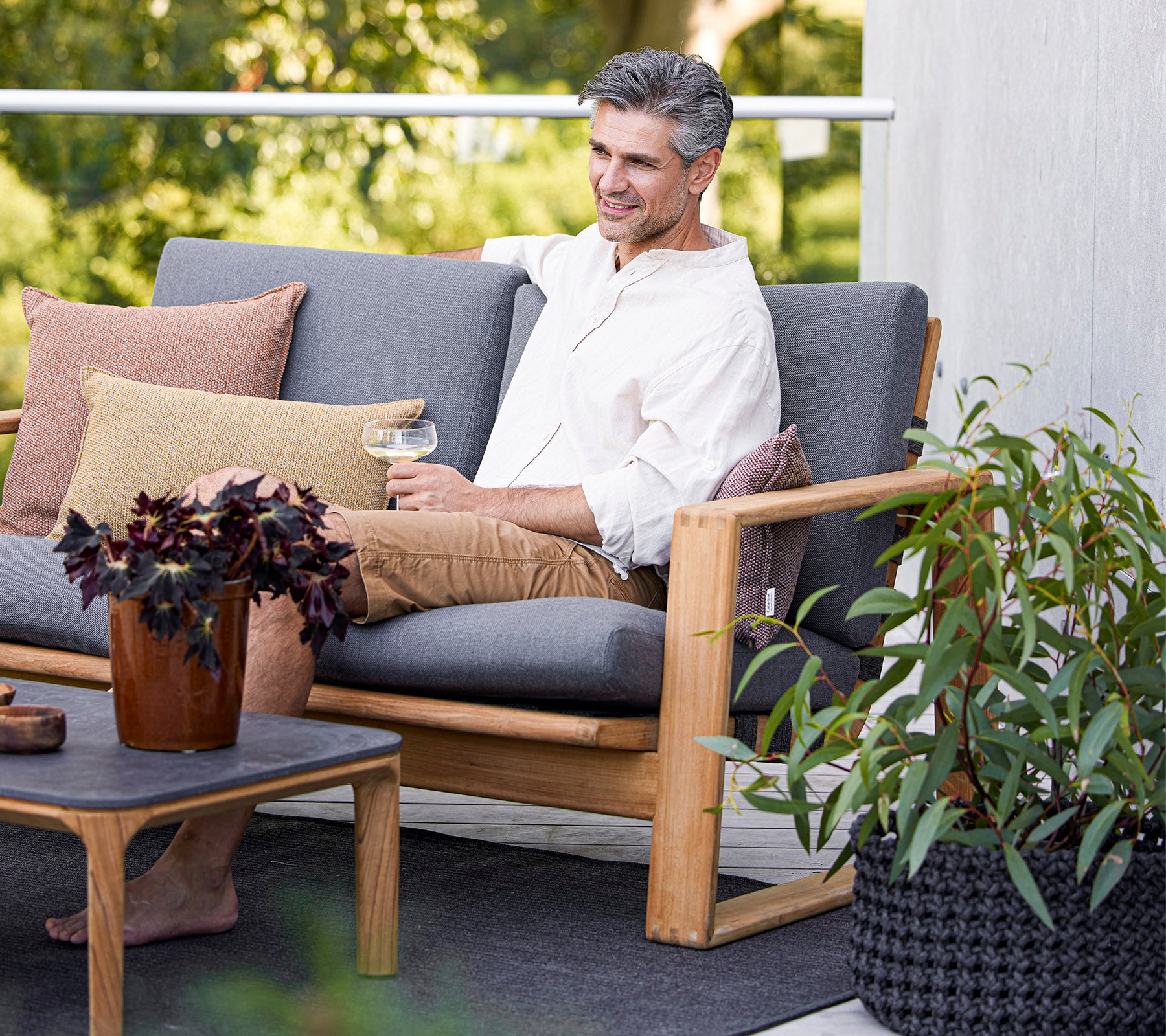 Un homme déguste une boisson tout en étant assis sur un canapé confortable, entouré de plantes et de coussins décoratifs.