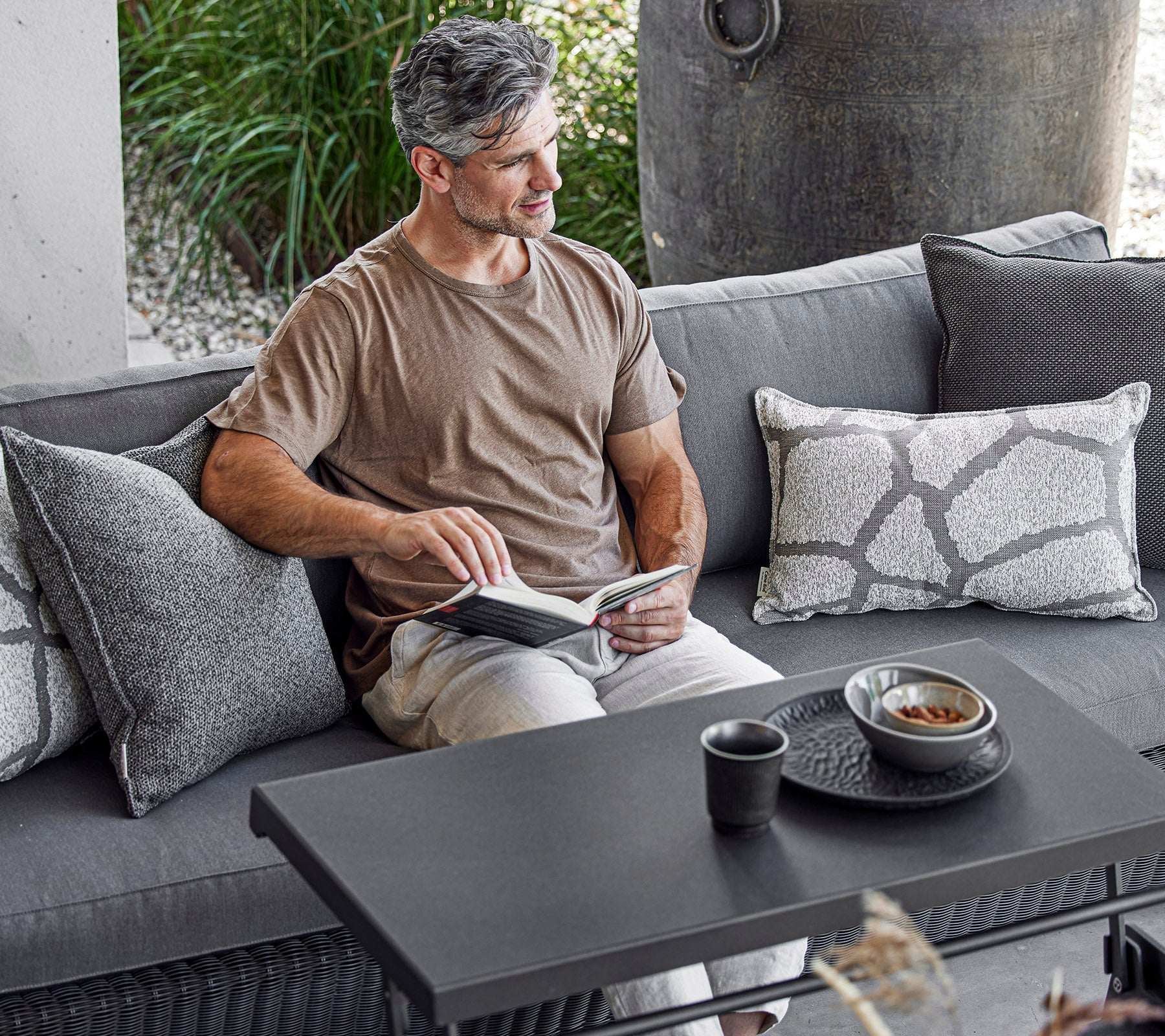 Un homme est assis confortablement sur un canapé, lisant un livre avec une tasse et un bol sur une table basse à côté de lui, entouré de coussins décoratifs.
